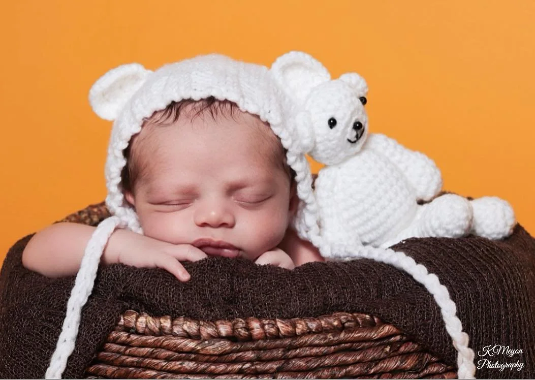 A sleeping baby wearing a white knit hat with bear ears, resting on a brown textured blanket, with a white crocheted teddy bear beside them, against a bright orange background.