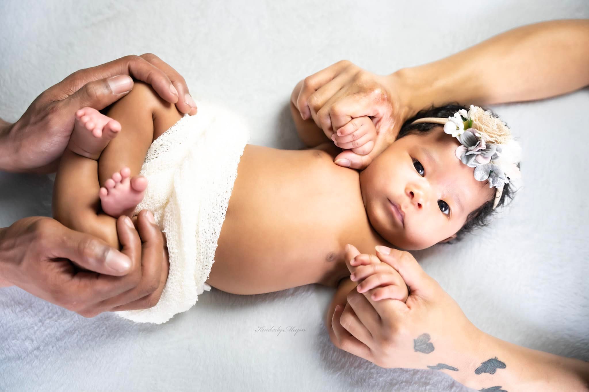 A baby girl with a flower headband lying on her side while adults hold her arm and hand gently.
