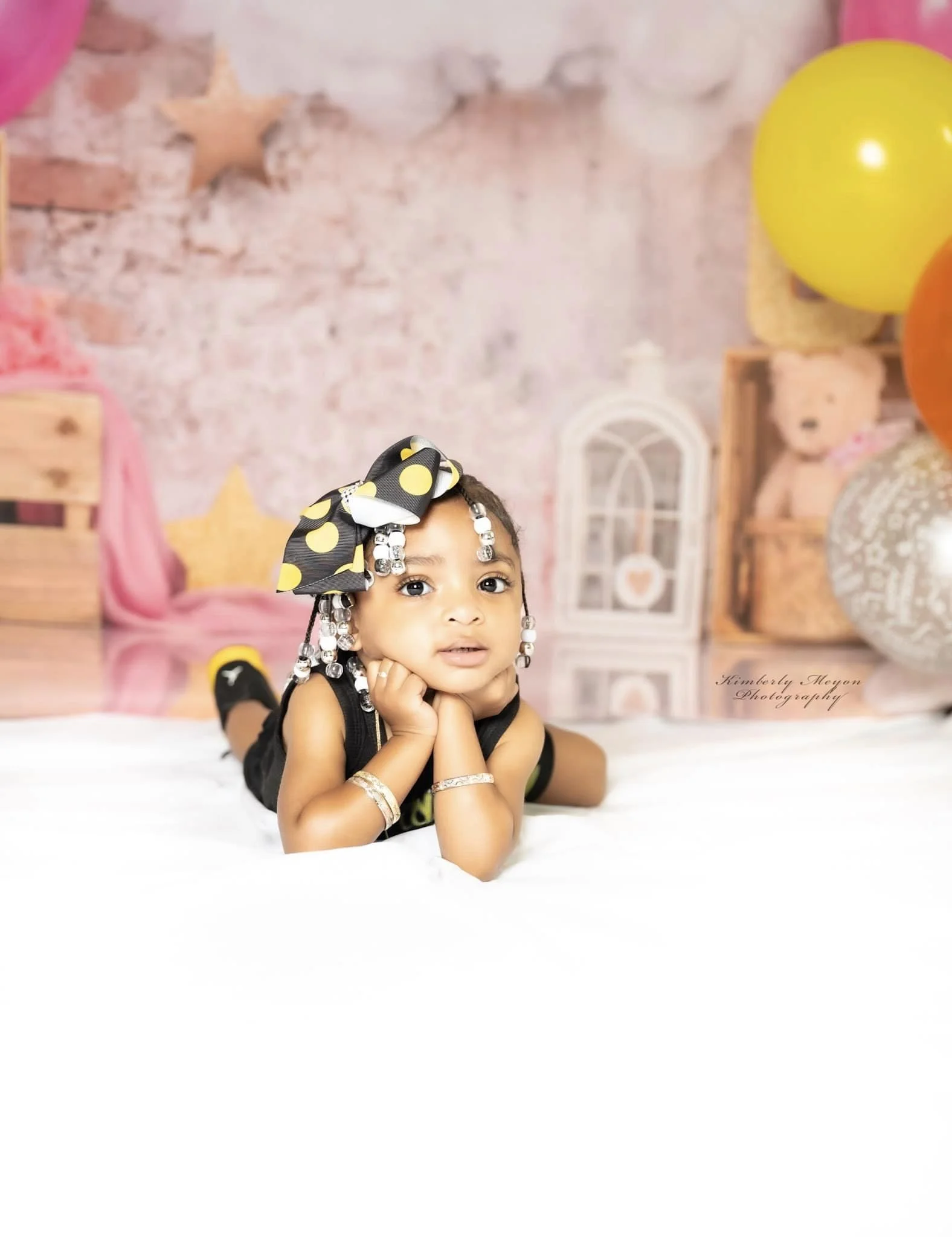 A young girl with braided hair and decorative beads, wearing a polka dot bow, lying on her stomach with her chin resting on her hands, in a pastel-colored room with balloons and teddy bears.