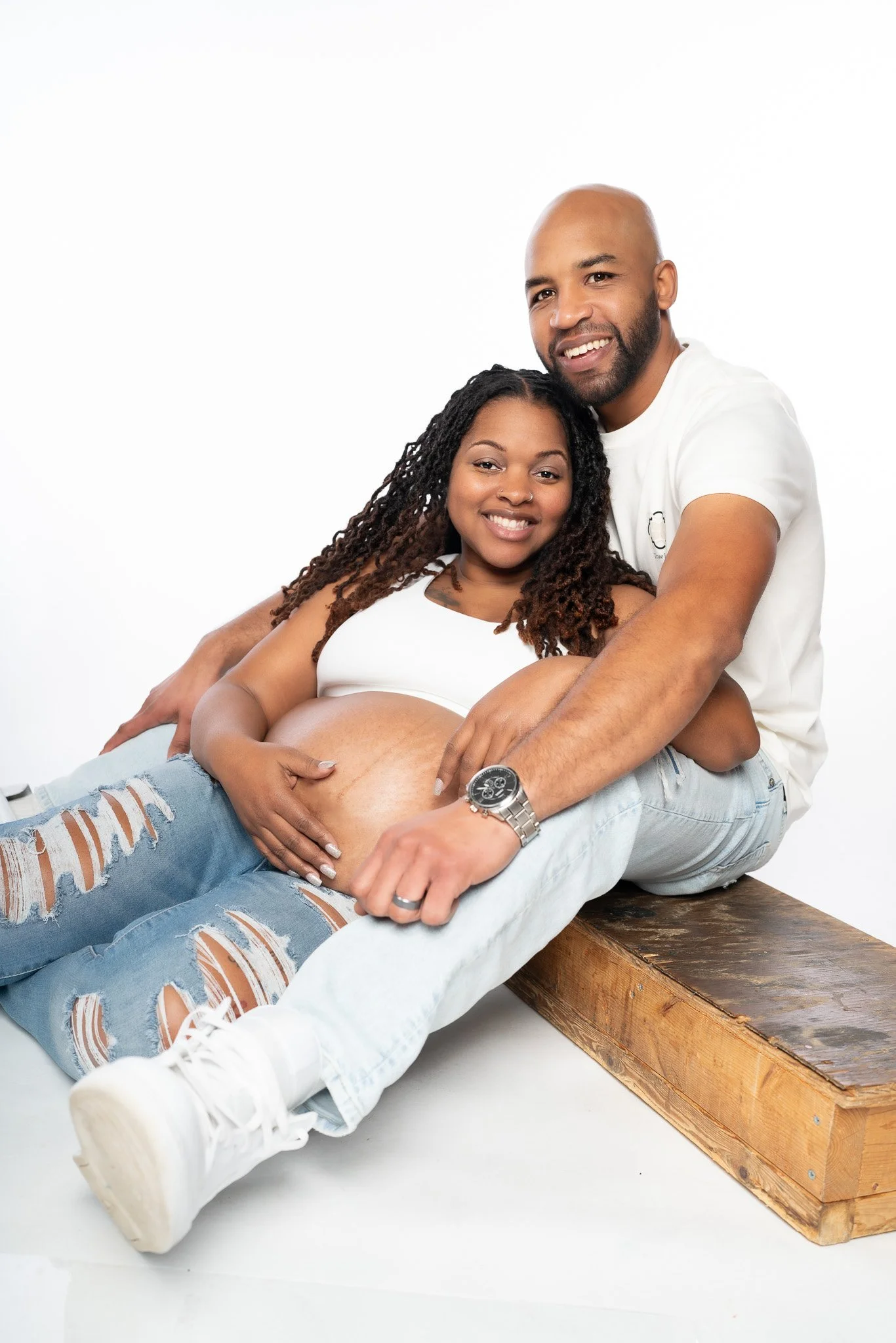 A joyful, pregnant woman with long curly hair lying on a wooden bench, with her partner hugging her from behind, both smiling at the camera against a plain white background.