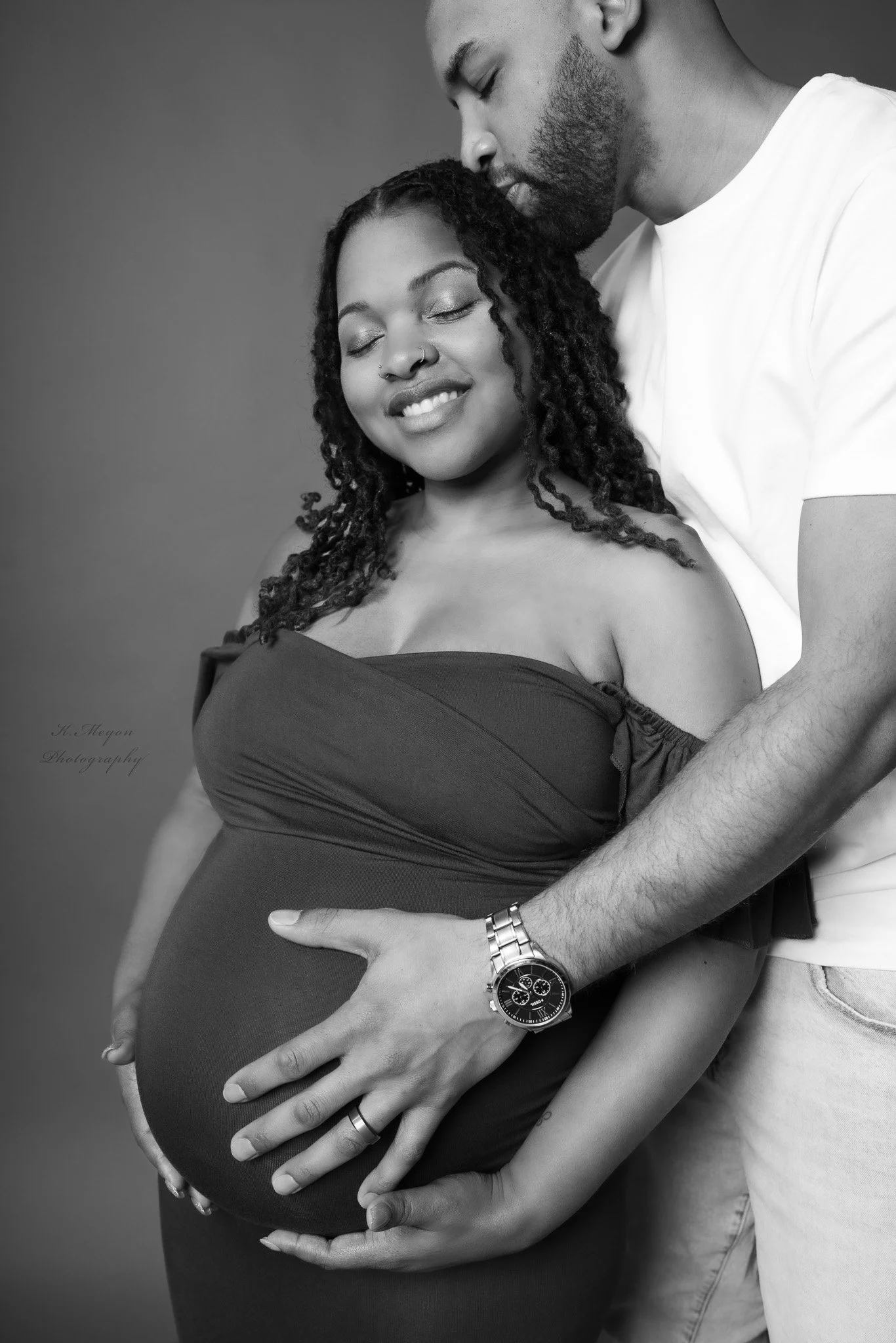 A pregnant woman with curly hair and a nose piercing, smiling with her eyes closed, is being embraced from behind by a man. The man, with a beard and wearing a white t-shirt, is resting his head gently on the woman's, with his eyes closed. Both are touching her pregnant belly and appear to be sharing a tender moment.