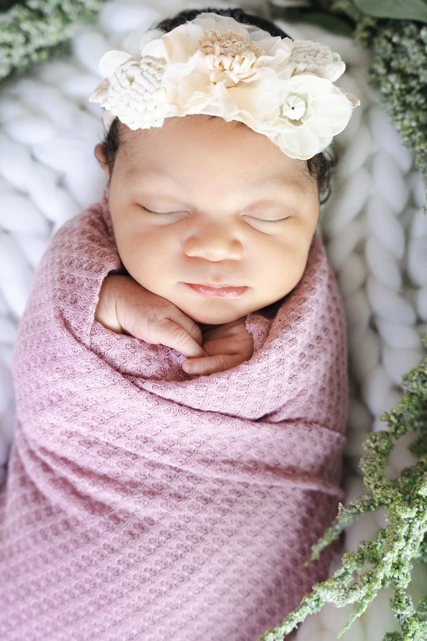 Close-up of a sleeping infant wearing a pink textured blanket and a floral headband, surrounded by greenery.