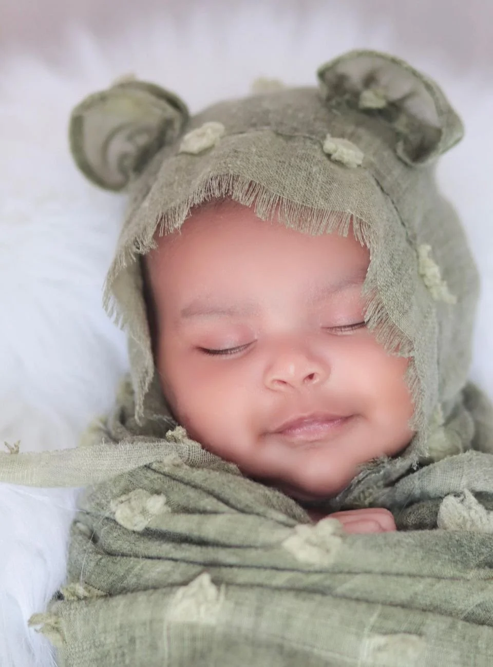 A sleeping baby dressed in a green, furry bear-themed hooded outfit with bear ears and frayed edges, resting with eyes closed.