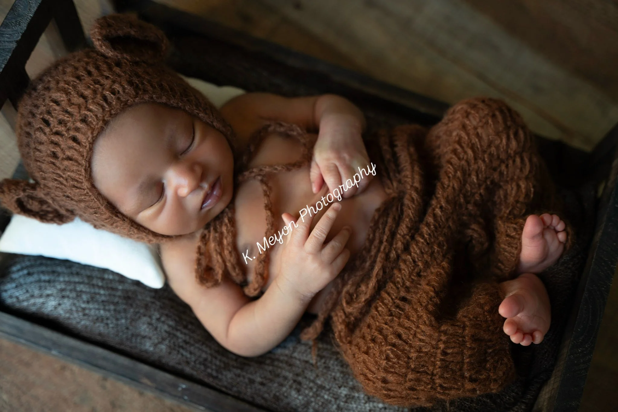 A young child sleeping peacefully wrapped in a cozy, brown knitted blanket with a matching knitted hood, resting on a cushioned surface.