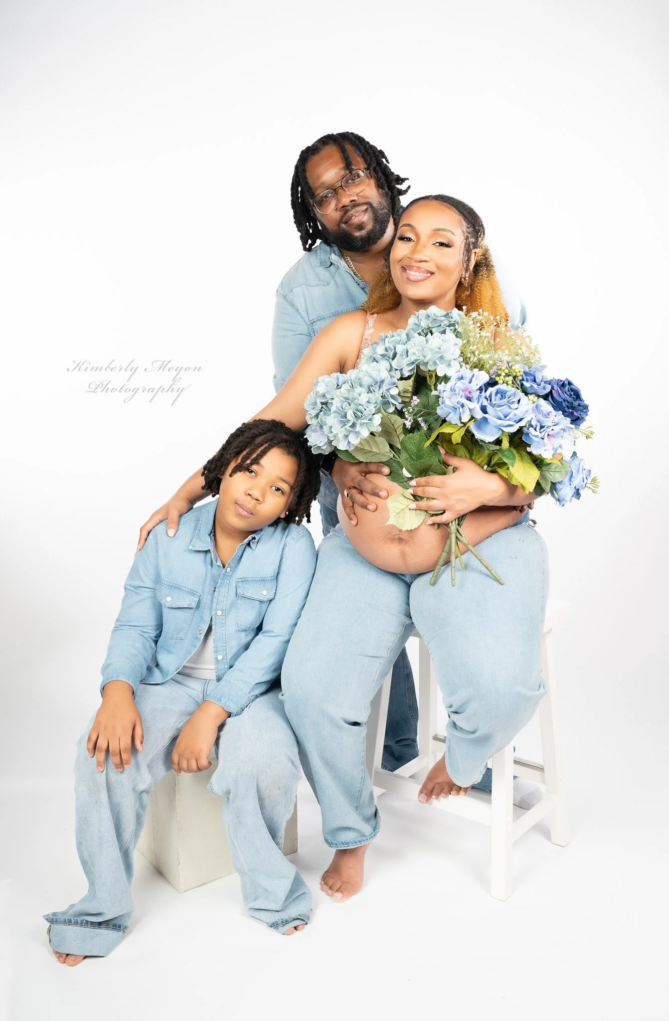 A family of four four posing for a studio portrait against a white background. The woman is sitting on a stool, holding a large bouquet of blue and purple flowers, and smiling at the camera. She has curly hair with blonde tips and is wearing a light denim outfit. Behind her, a man with glasses and dreadlocks is leaning in, also smiling. A young girl with dreadlocks and wearing a denim jacket is seated on a white block in front of the woman, looking slightly to the side.