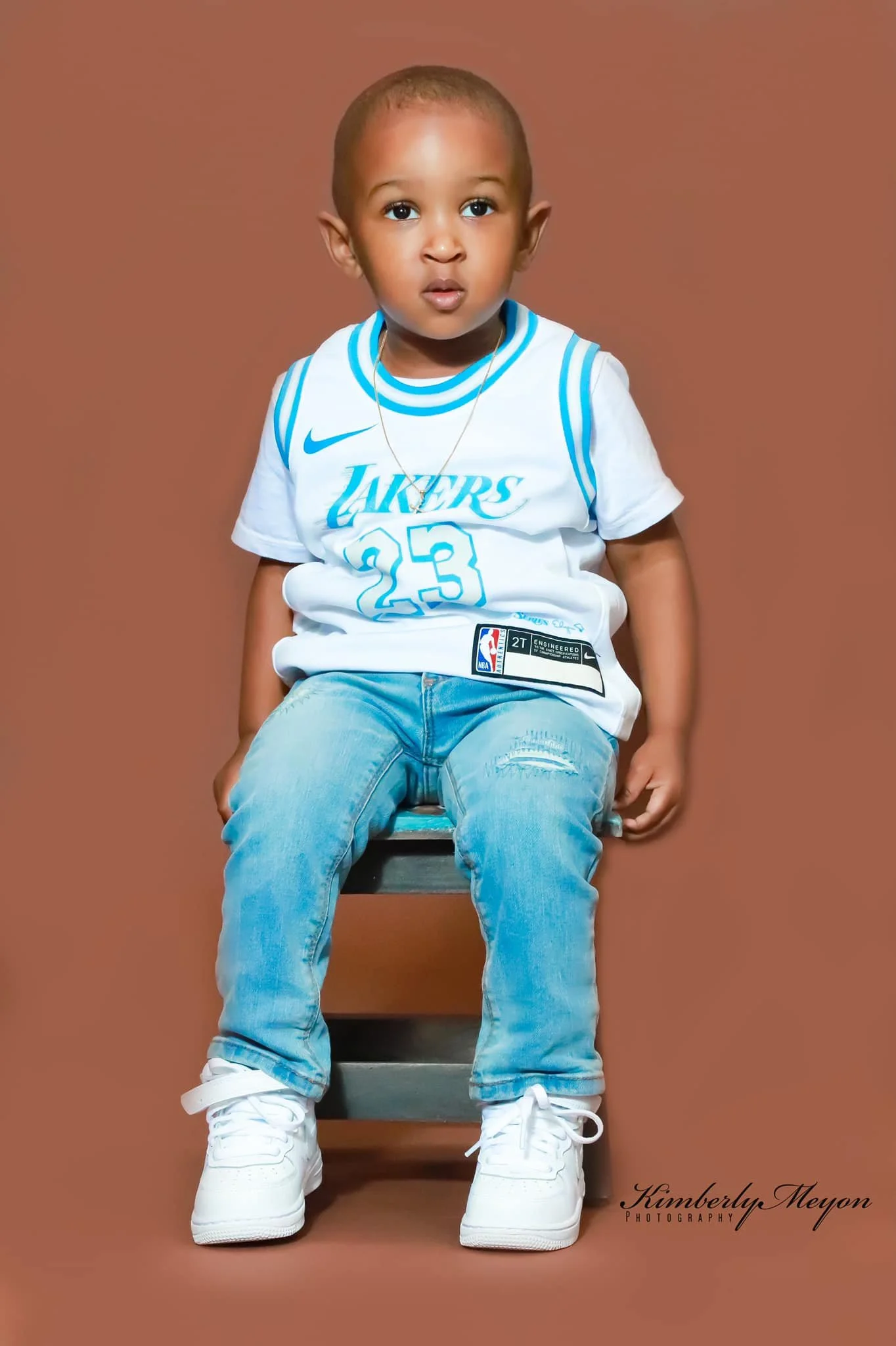 Young boy wearing a white Lakers basketball jersey with the number 23, ripped light blue jeans, white sneakers, a gold chain, seated on a staircase against a brown background.