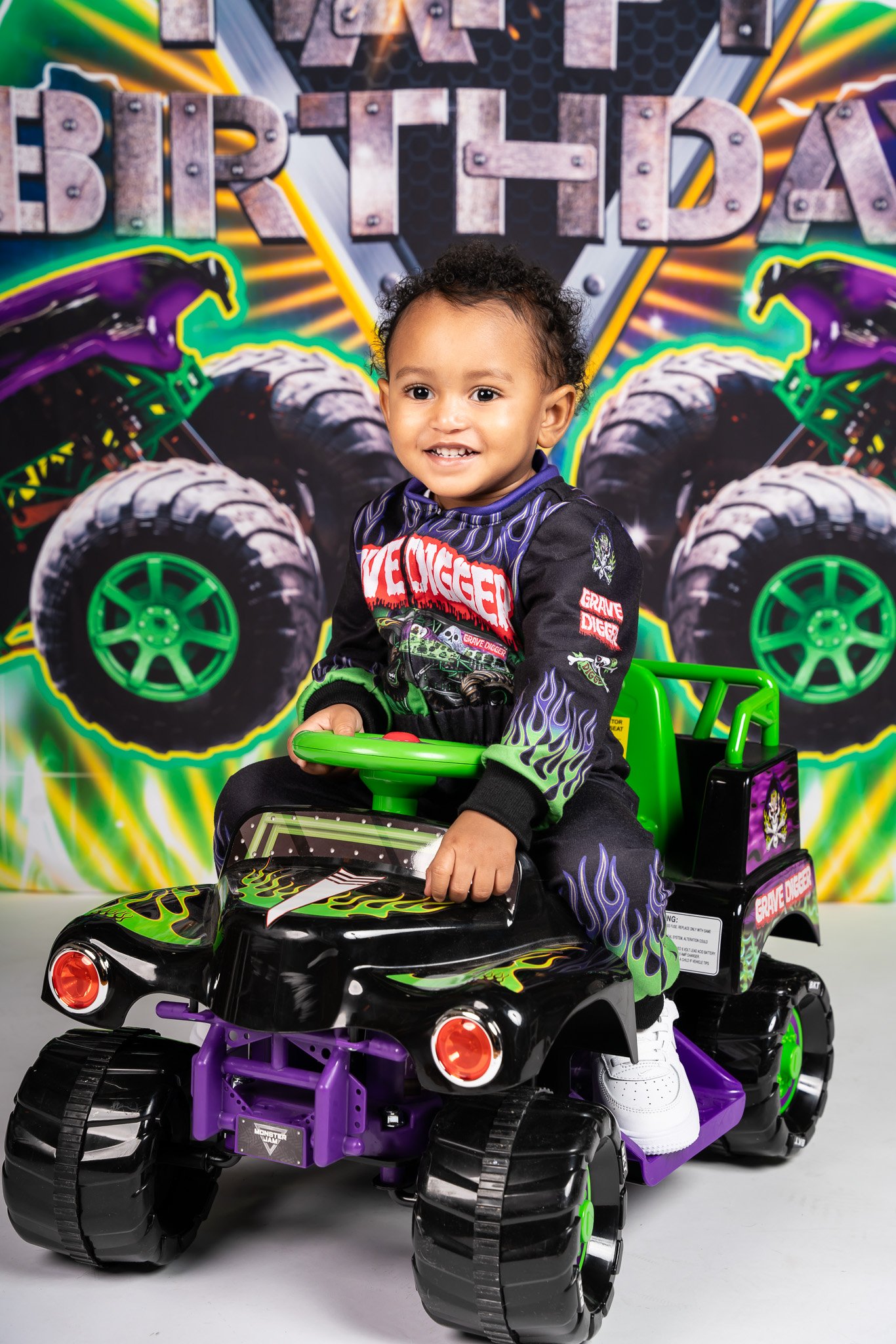 Young child smiling and sitting in a black and purple toy car with neon green accents, in front of a colorful background with large metallic and neon lettering.