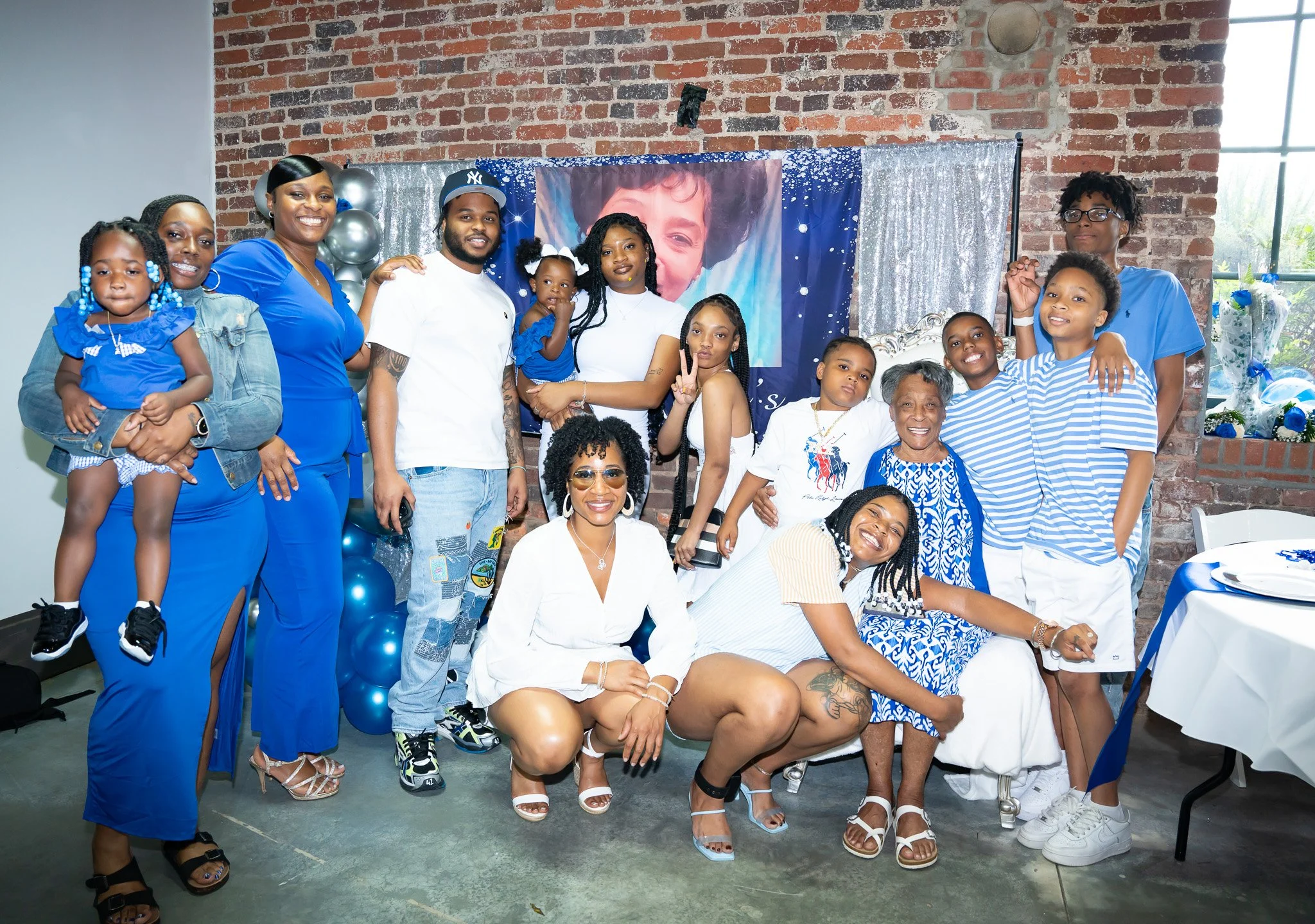 Family group photo at a celebration in front of a portrait of a smiling person. Several people, including children and adults, are dressed in blue and white, smiling and posing for the camera with festive decorations, balloons, and a table with gifts and flowers.