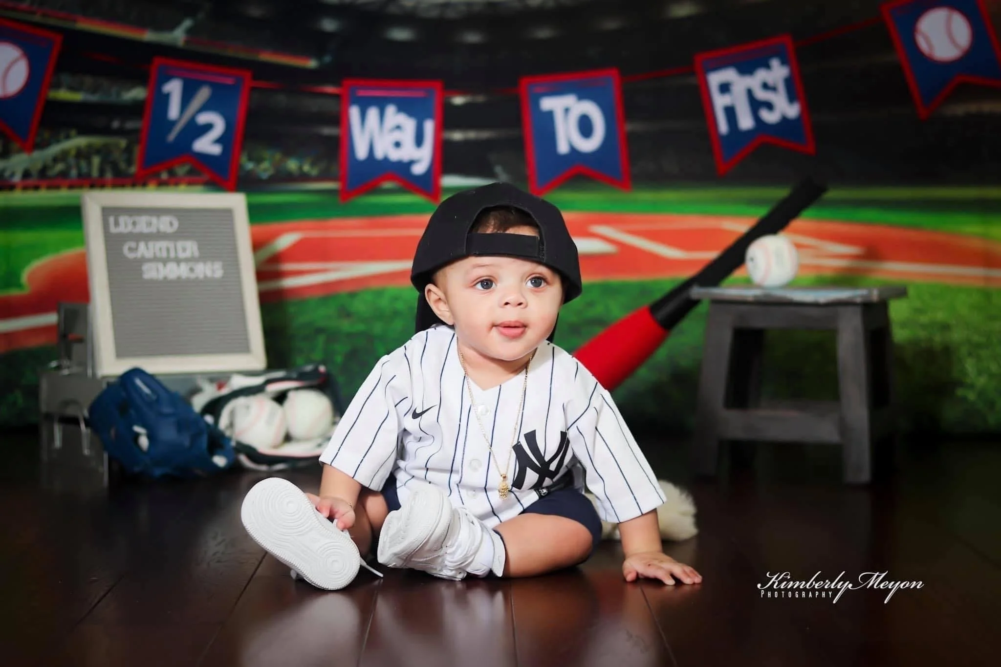 Young child dressed in a baseball jersey and baseball cap sitting on the floor with a baseball glove and sneakers nearby, in front of a baseball-themed mural with banners that read '1/2 May 10' and a sign that says 'Used Canner Space'.
