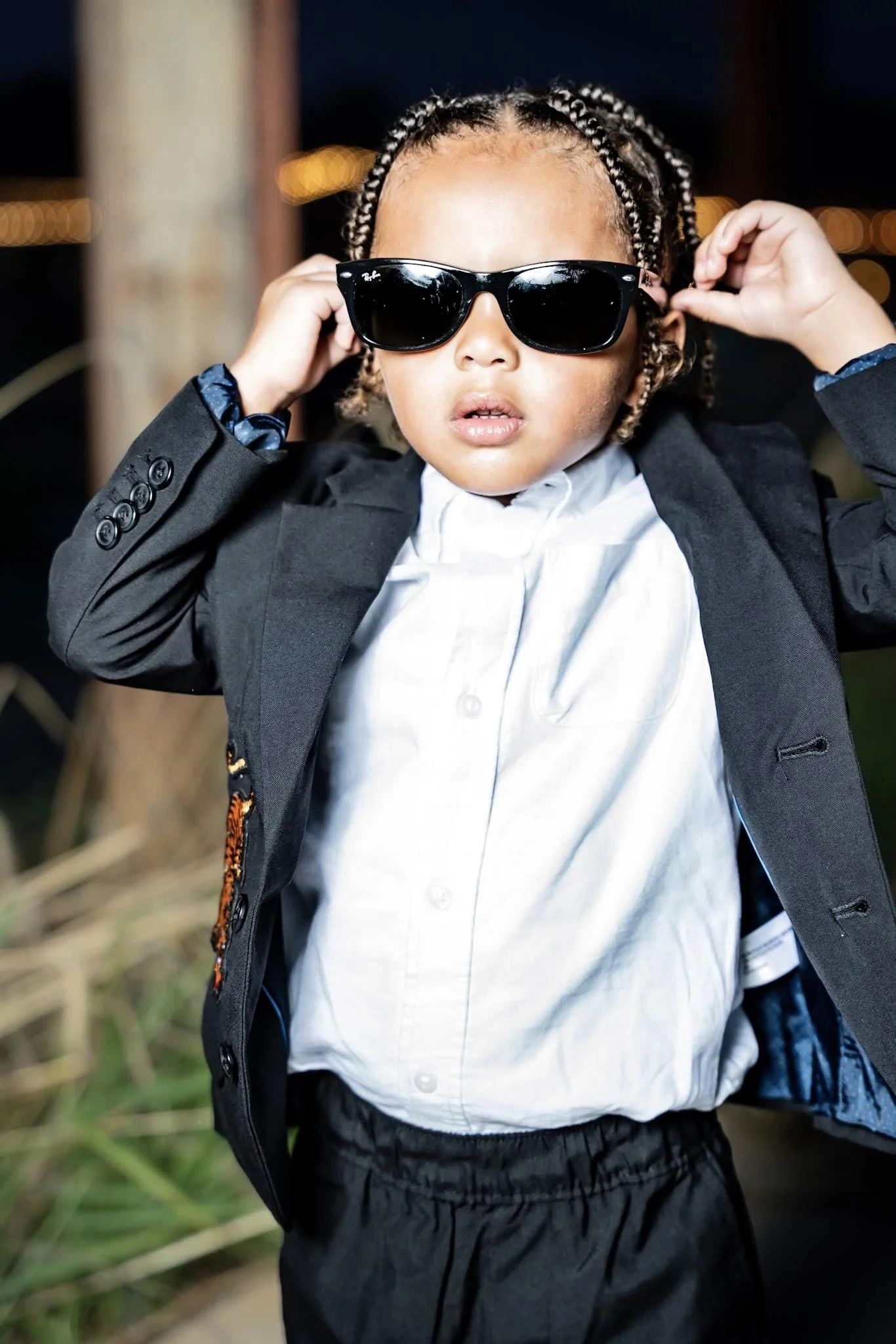 A young boy wearing dark sunglasses, a black blazer, a white shirt, and black shorts. He is adjusting his sunglasses with both hands and standing outdoors at night with blurred lights and natural elements in the background.