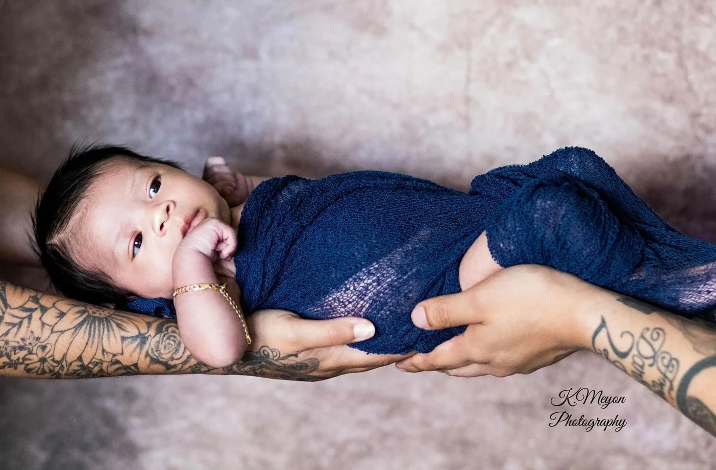 A newborn baby wrapped in a blue cloth being held by two tattooed hands, one with a bracelet, against a neutral background.