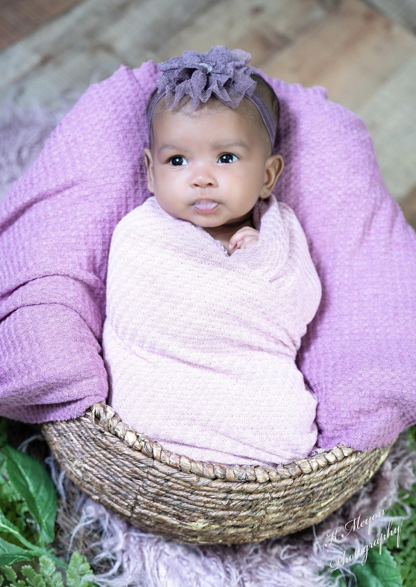 A baby wrapped in a light pink cloth, lying in a woven basket with a purple headband with a bow, outdoors on grass and a stone pathway.