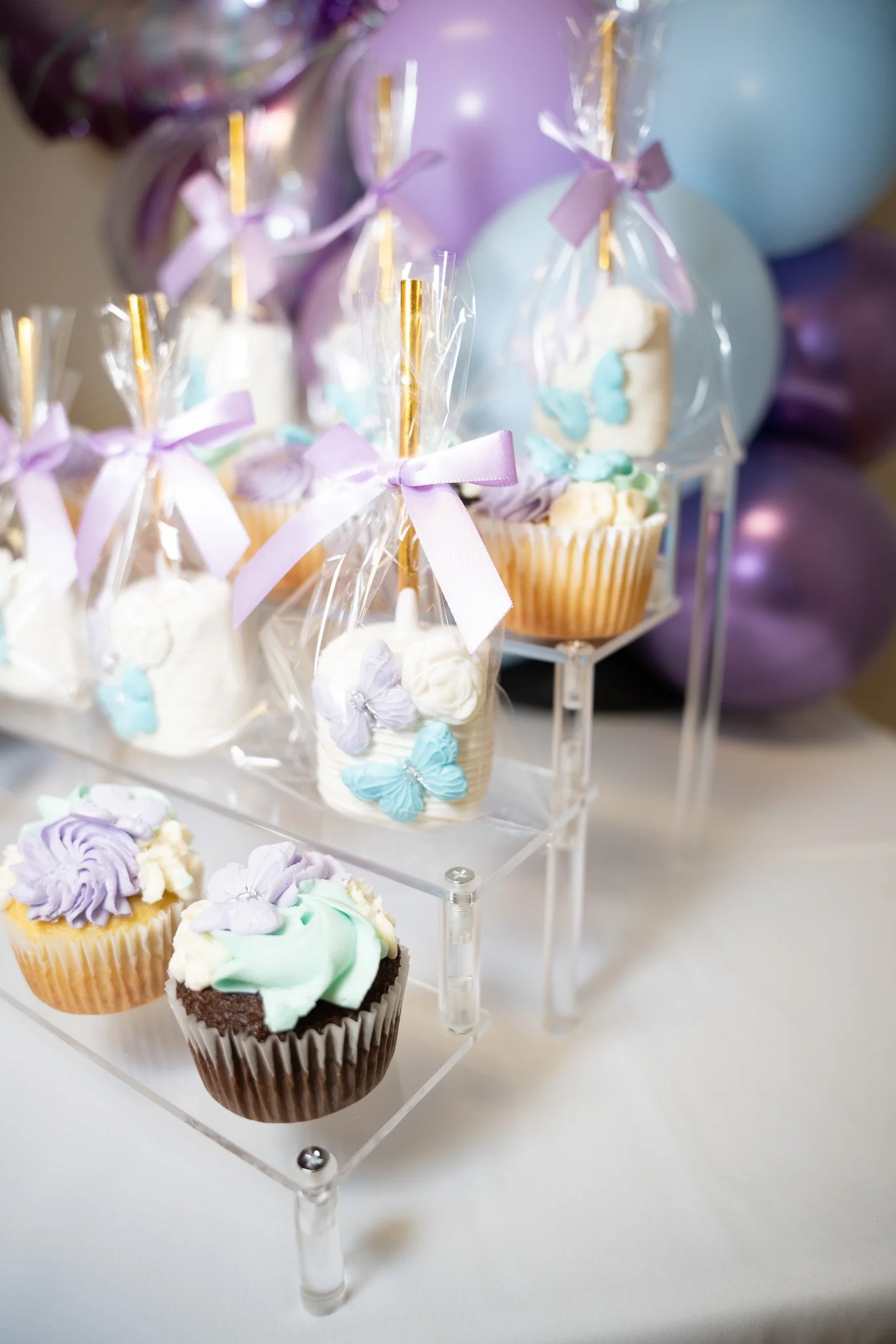 Assorted cupcakes decorated with pastel-colored frosting and butterfly-shaped embellishments, displayed on a clear acrylic stand with purple balloons in the background.