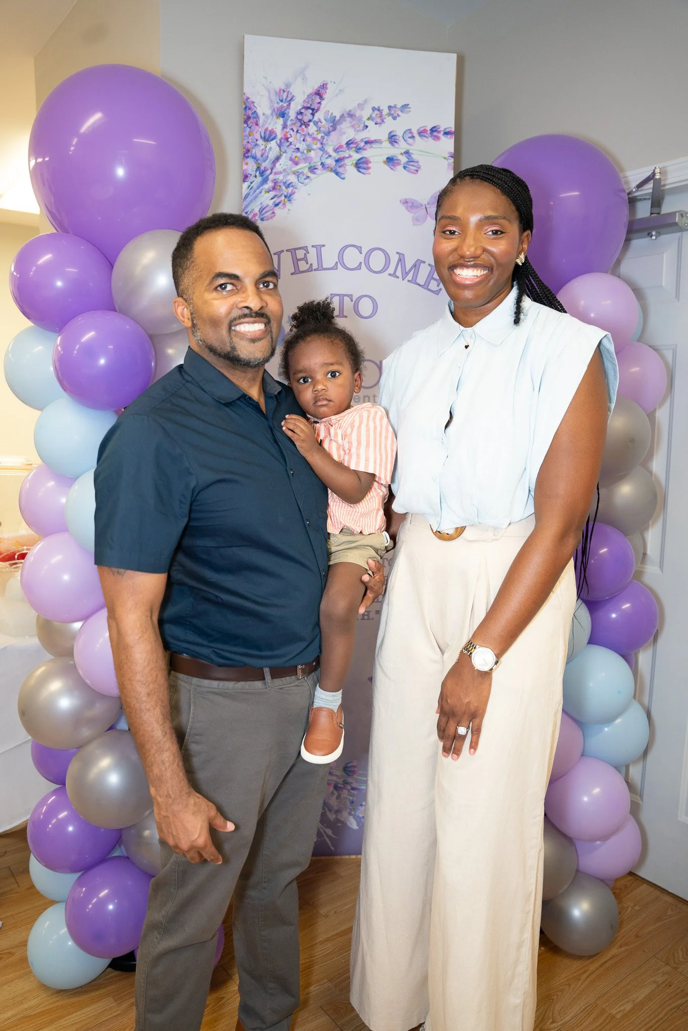 A family of three standing in front of a welcome sign decorated with purple, white, and silver balloons, with a purple and white floral artwork behind them. The man is wearing a navy blue shirt and gray pants, the woman is wearing a white sleeveless top and white pants, and the young girl is wearing an orange striped outfit.