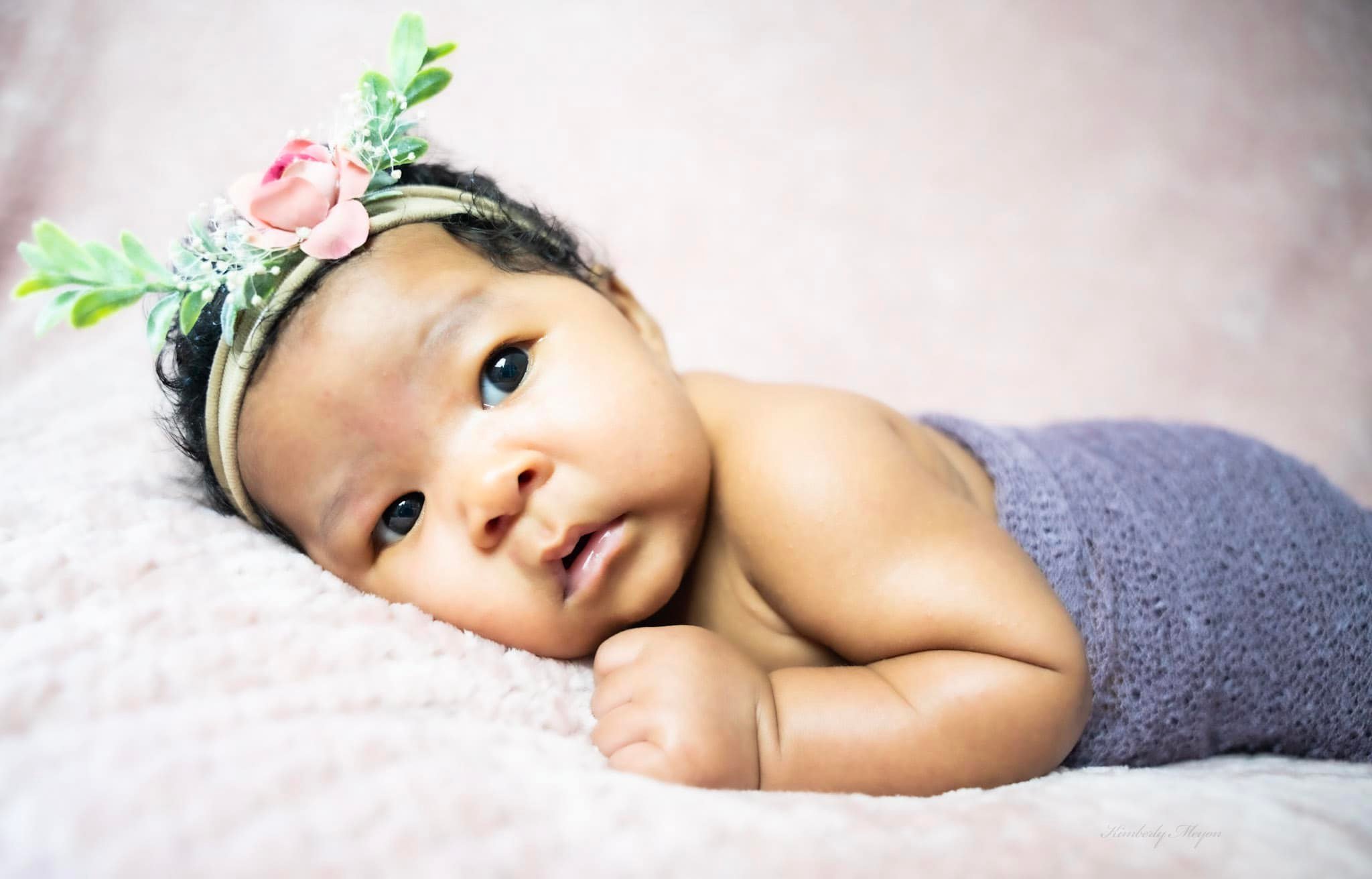 Close-up of a baby lying on a soft surface, wearing a floral headband, looking at the camera with wide eyes.