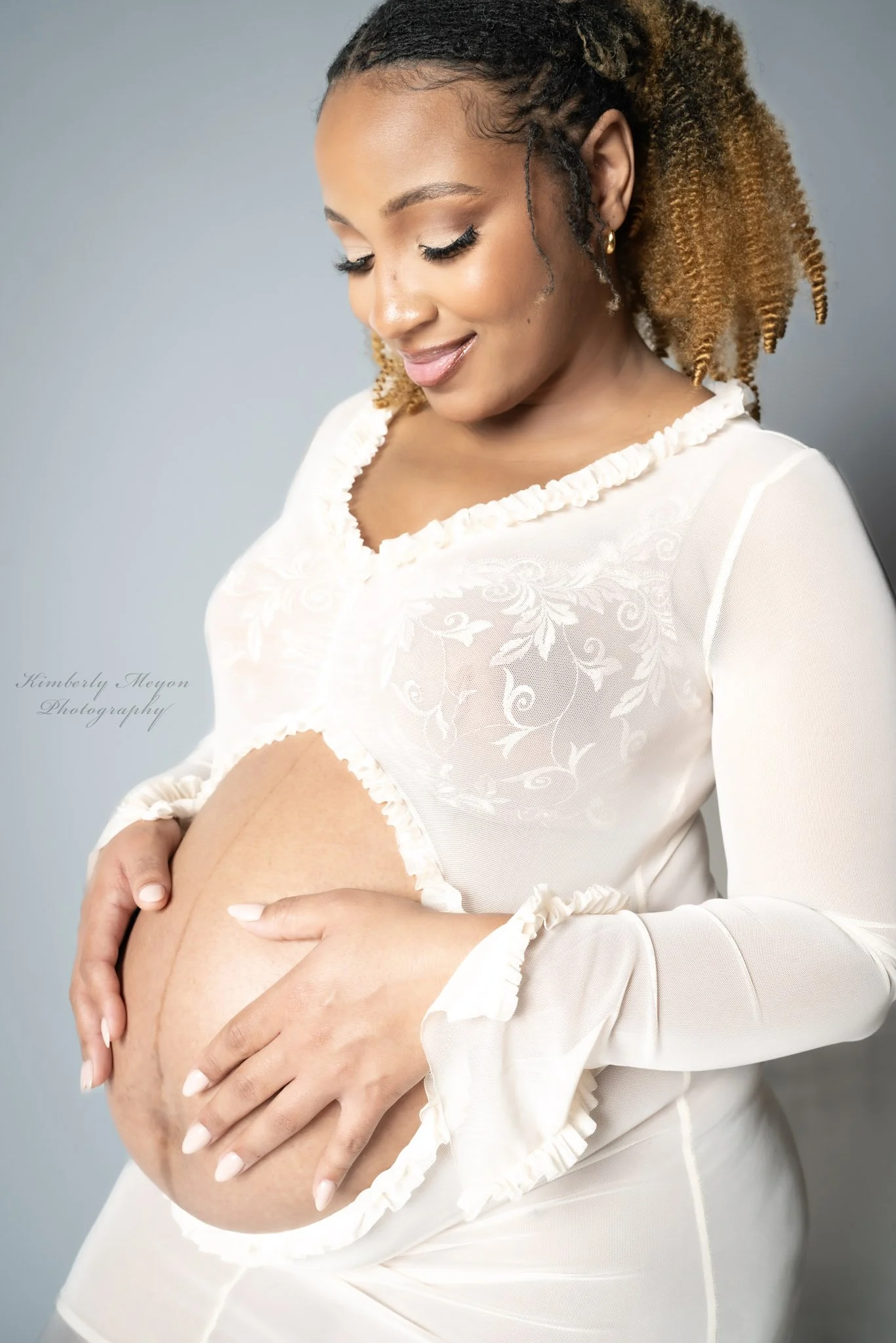 A pregnant woman with curly black and blonde hair gently holding her belly, smiling softly with her eyes closed, wearing a white, long-sleeved, embroidered blouse, against a plain gray background.
