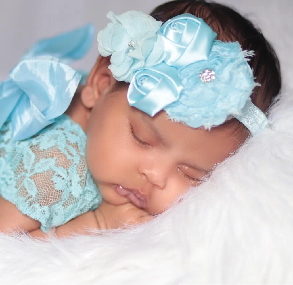 Close-up of a sleeping baby girl with a blue floral headband and lace dress, resting on a soft white blanket.