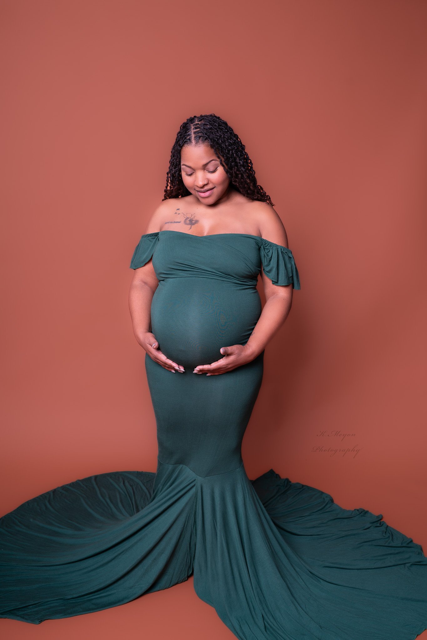 A pregnant woman with curly hair wearing a teal off-shoulder dress, standing against a plain brown background, gently holding her belly with both hands, with her eyes closed and a peaceful expression.