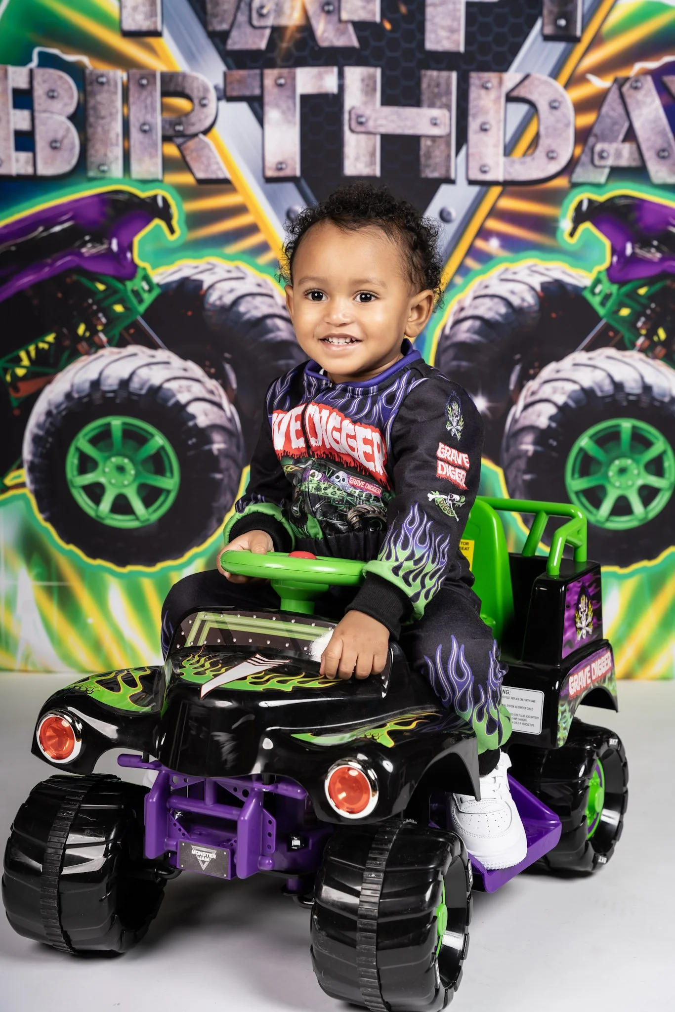 A young boy with curly hair smiling while sitting on a black and purple toy monster truck. The boy wears a black racing jersey with colorful flames and logos, and white sneakers. Behind him is a vibrant 'Monster Jam' themed backdrop with tire graphic