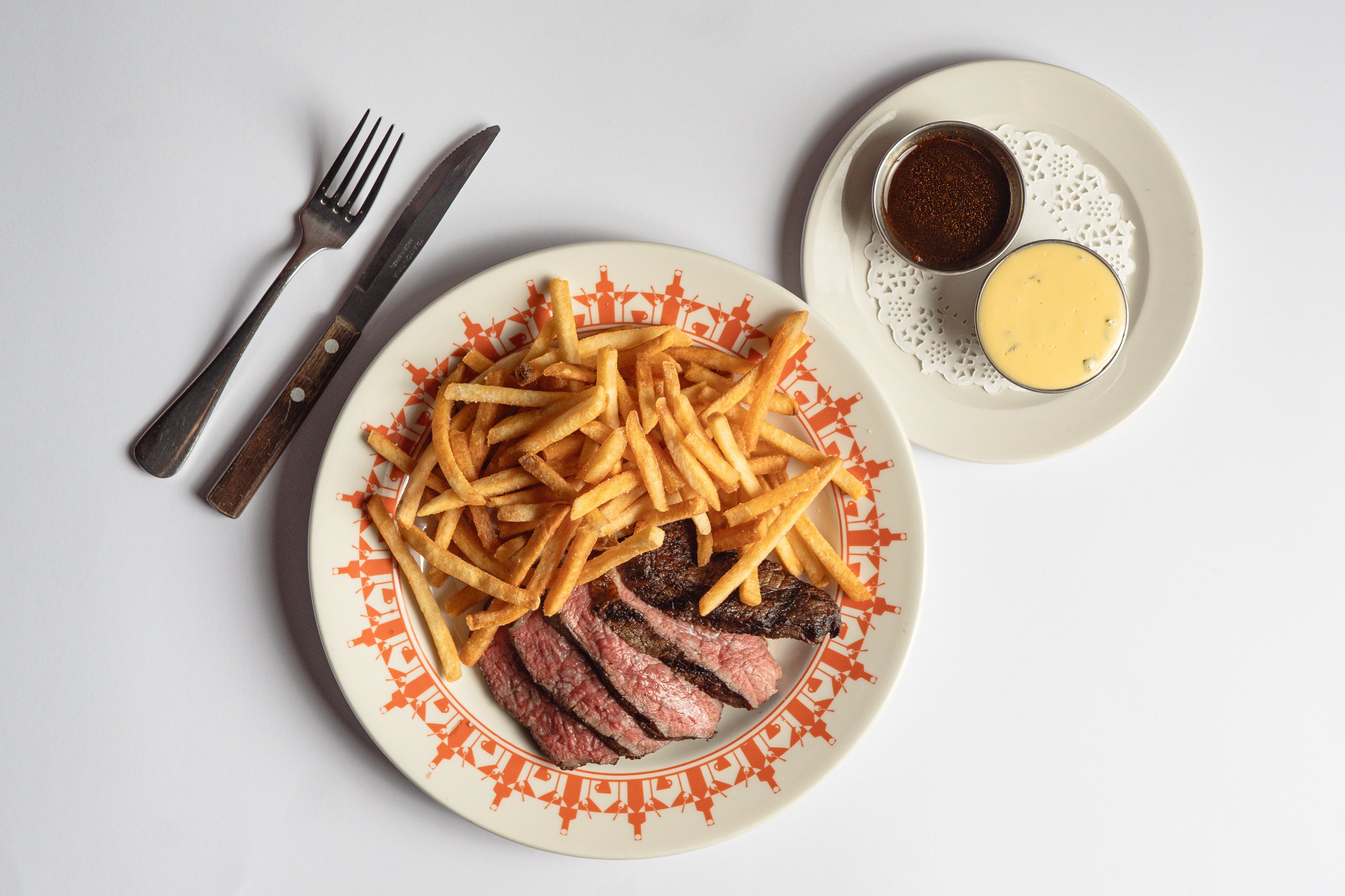 Plate of sliced steak and French fries with two dipping sauces on a side plate, with a fork and knife on a white background.