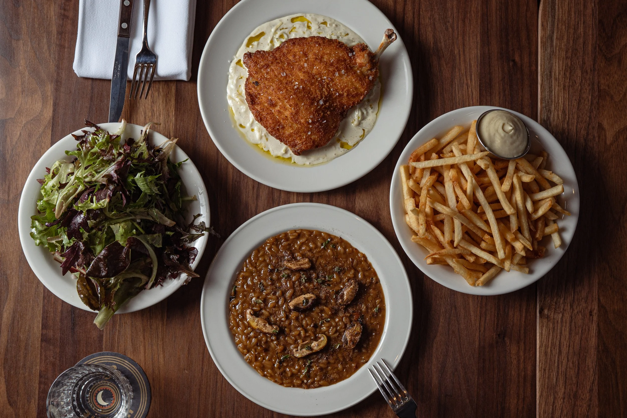 A top-down view of a meal including a salad, a plate of fried chicken with gravy, a plate of French fries with dipping sauce, and a bowl of baked beans on a wooden table.
