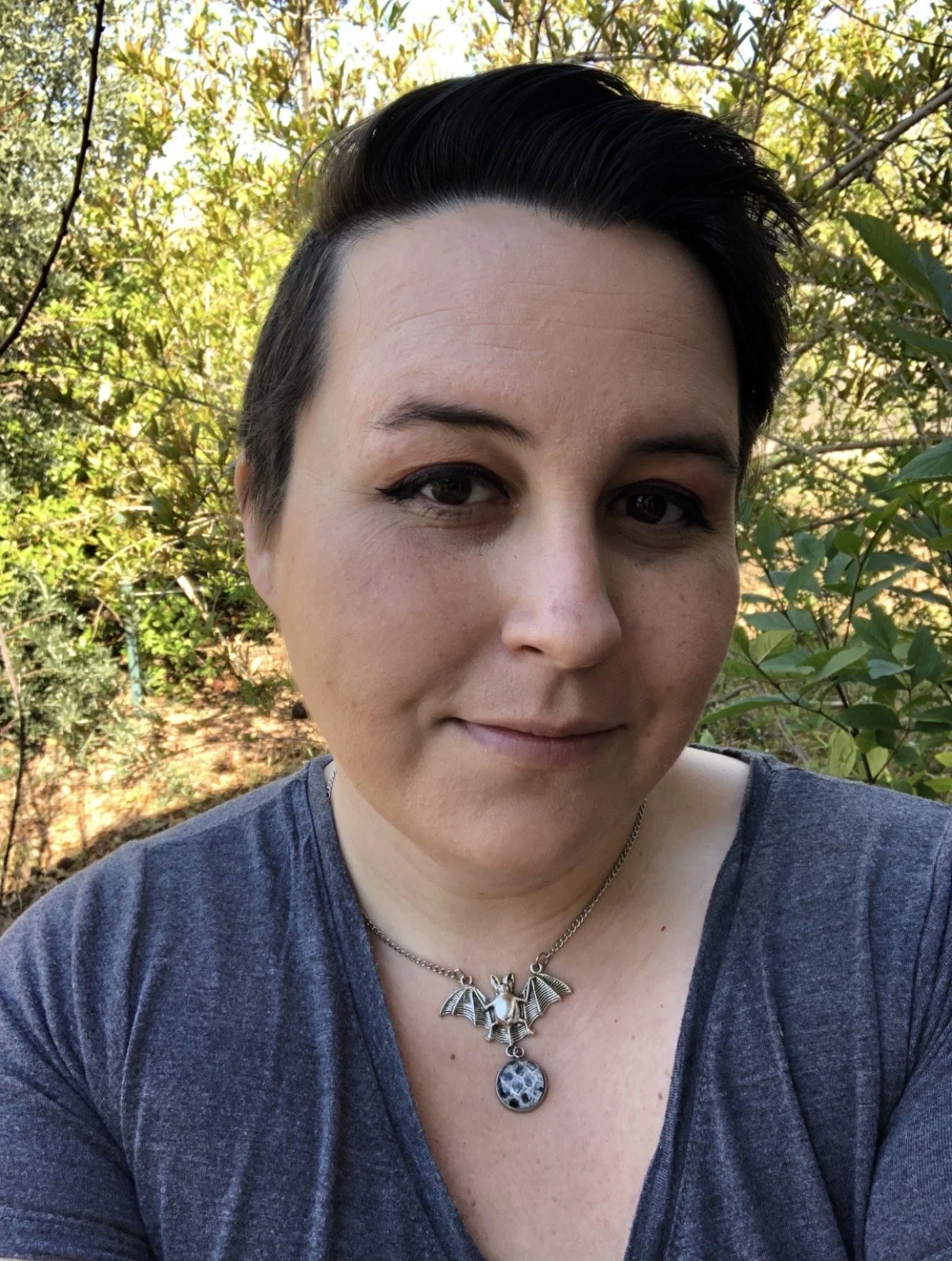 Close-up of a woman with short dark hair, wearing a gray shirt and a silver necklace with a bat-shaped pendant, outdoors with green foliage in the background.