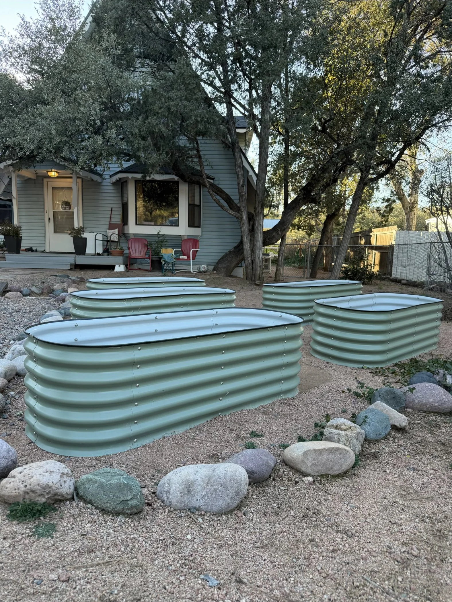 Decorative raised garden beds made of light green corrugated metal, arranged in a backyard with trees, rocks, and a house with a porch in the background.