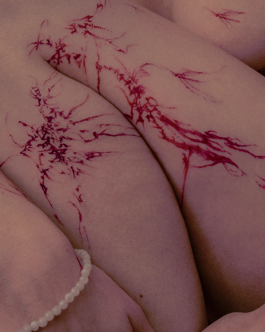 Close-up of a person's body with red ink sketches resembling tree branches and roots drawn on the skin, alongside a pearl bracelet.