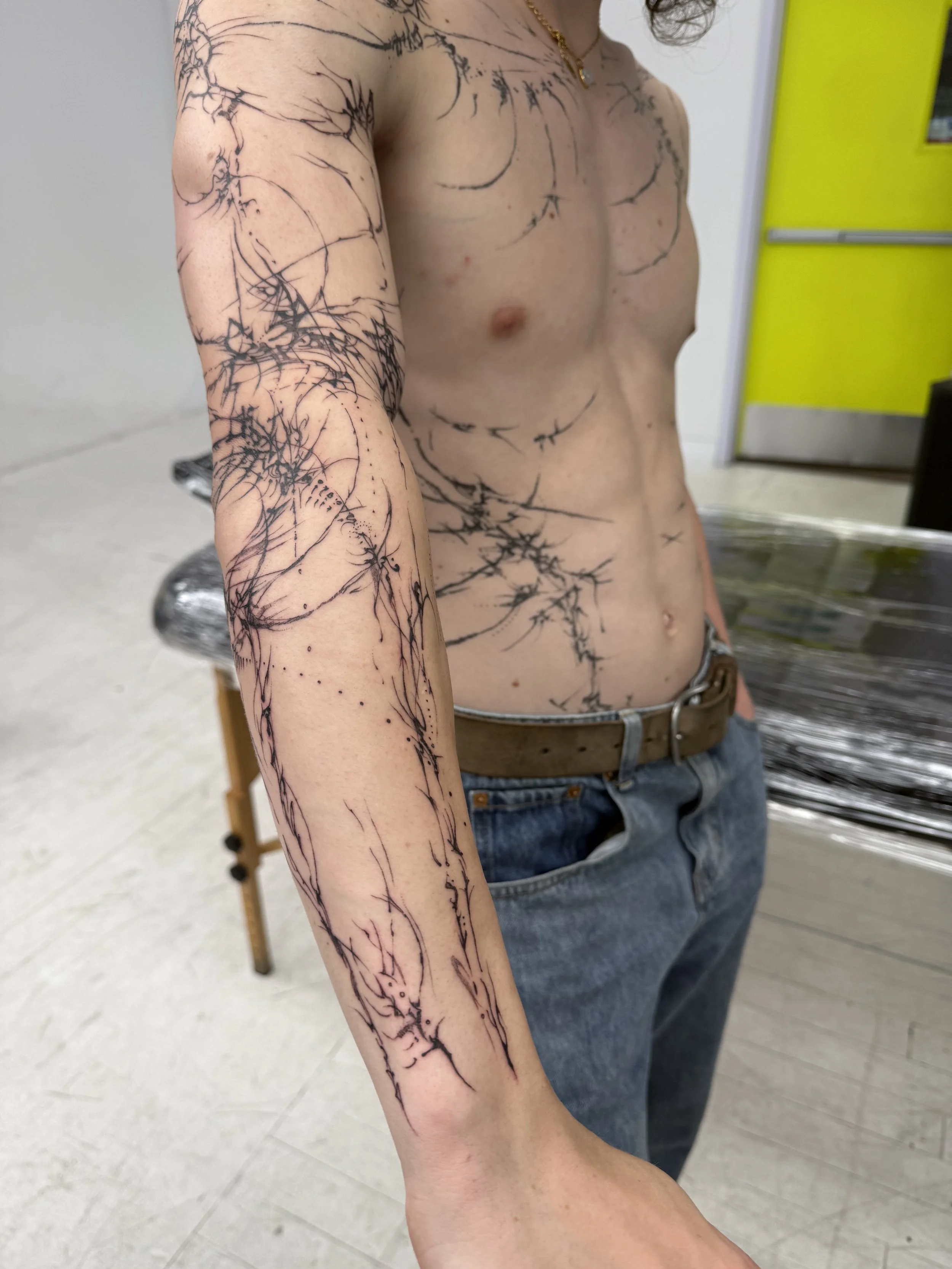 Person with freshly tattooed black ink abstract line design on arm and torso, shirtless, in a studio or shop with yellow door in background.