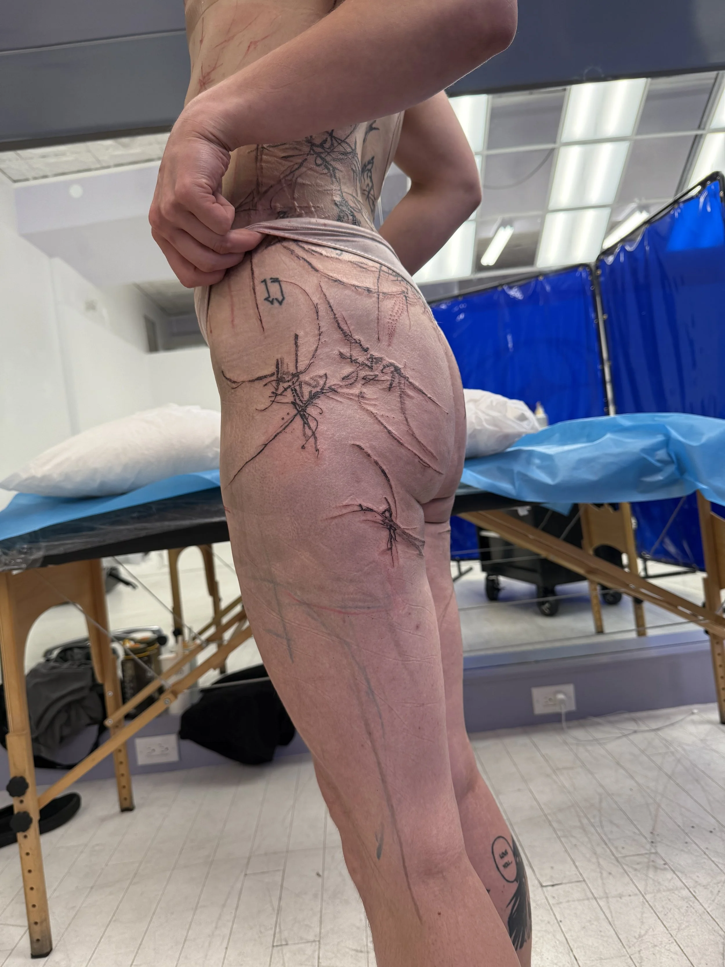 A person standing in a medical or tattoo studio, partially undressed, showing their side profile and tattoos on their body, with markings on their skin indicating a tattoo or medical procedure in progress.