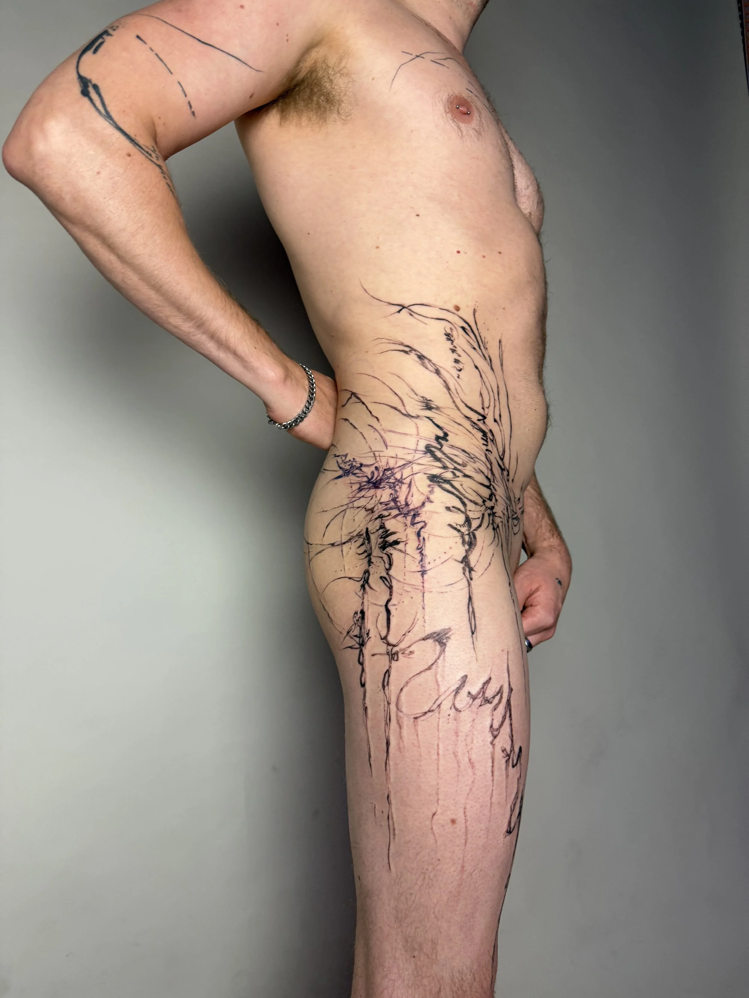 Side view of a shirtless man with tattoos on his arm, side, and thigh, standing against a plain wall.