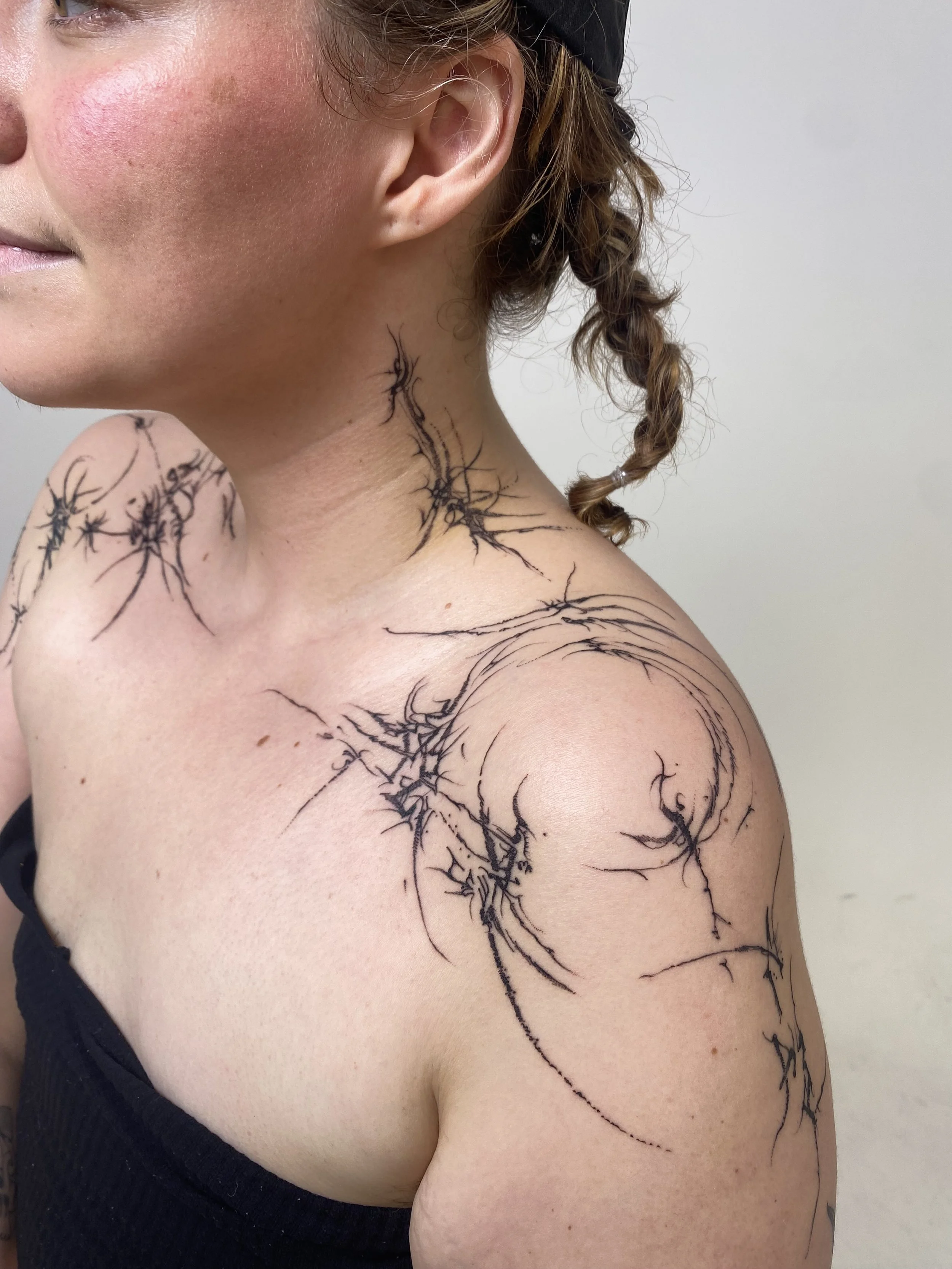 Close-up side view of a woman with short hair wearing a black tank top, showing her shoulder and neck covered in black ink tattoo designs resembling cracks or lightning patterns.