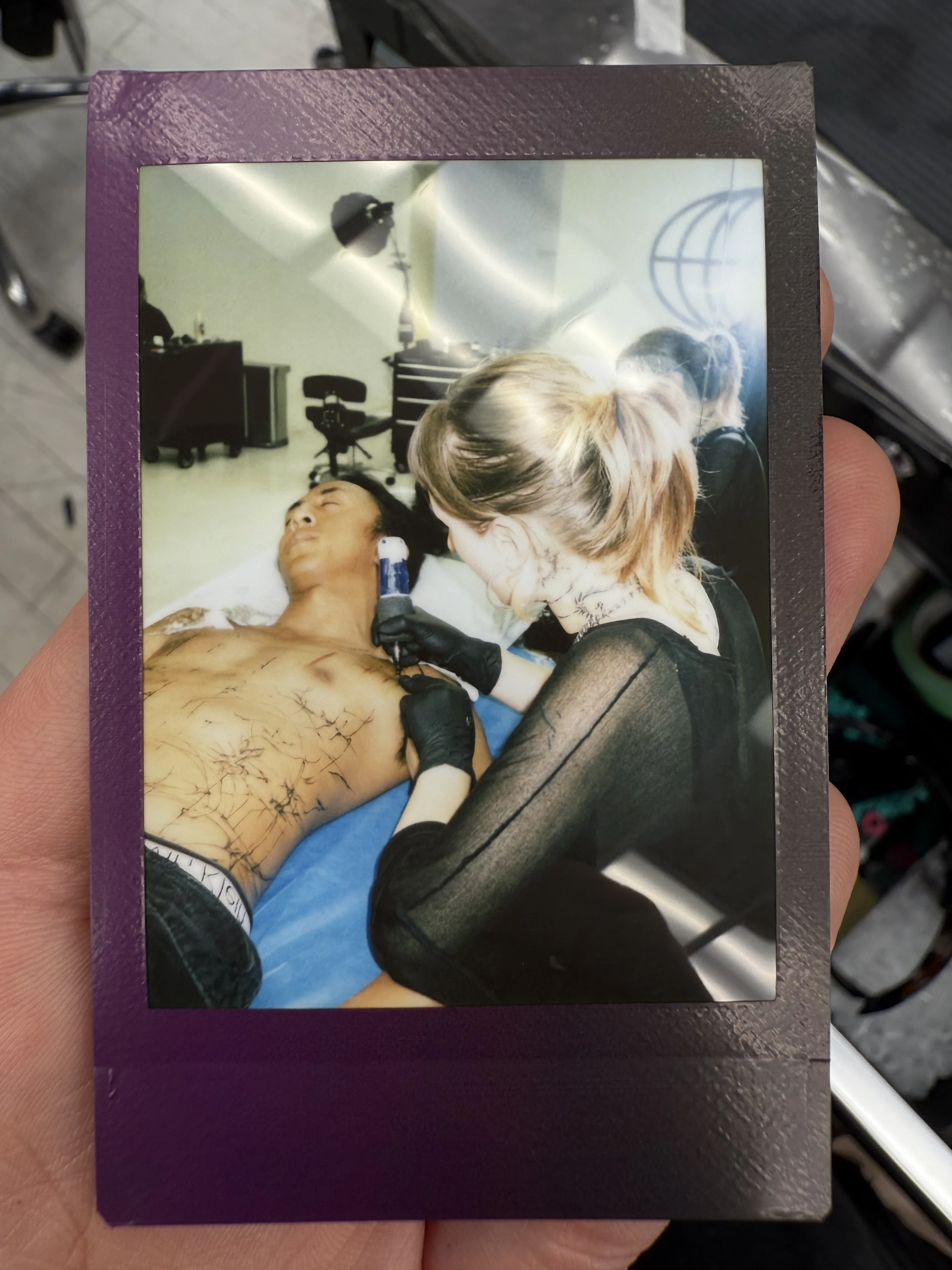 A woman is tattooing a man's chest with a device while he lays on an examination table, surrounded by tattoo equipment and a tattoo studio setting.