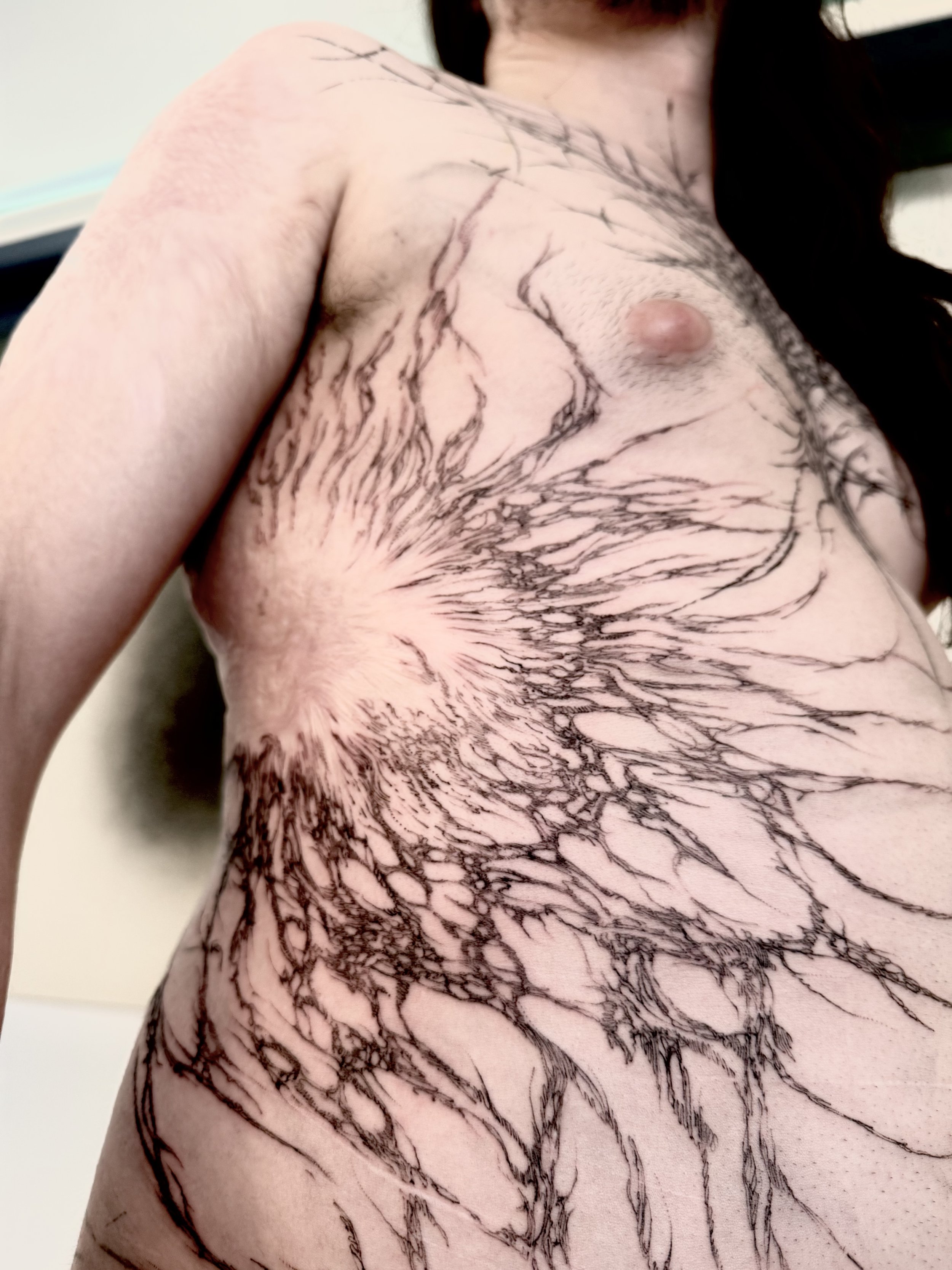 Close-up of a person's upper torso with a large, dark, branch-like tattoo covering the shoulder, chest, and side of the torso.