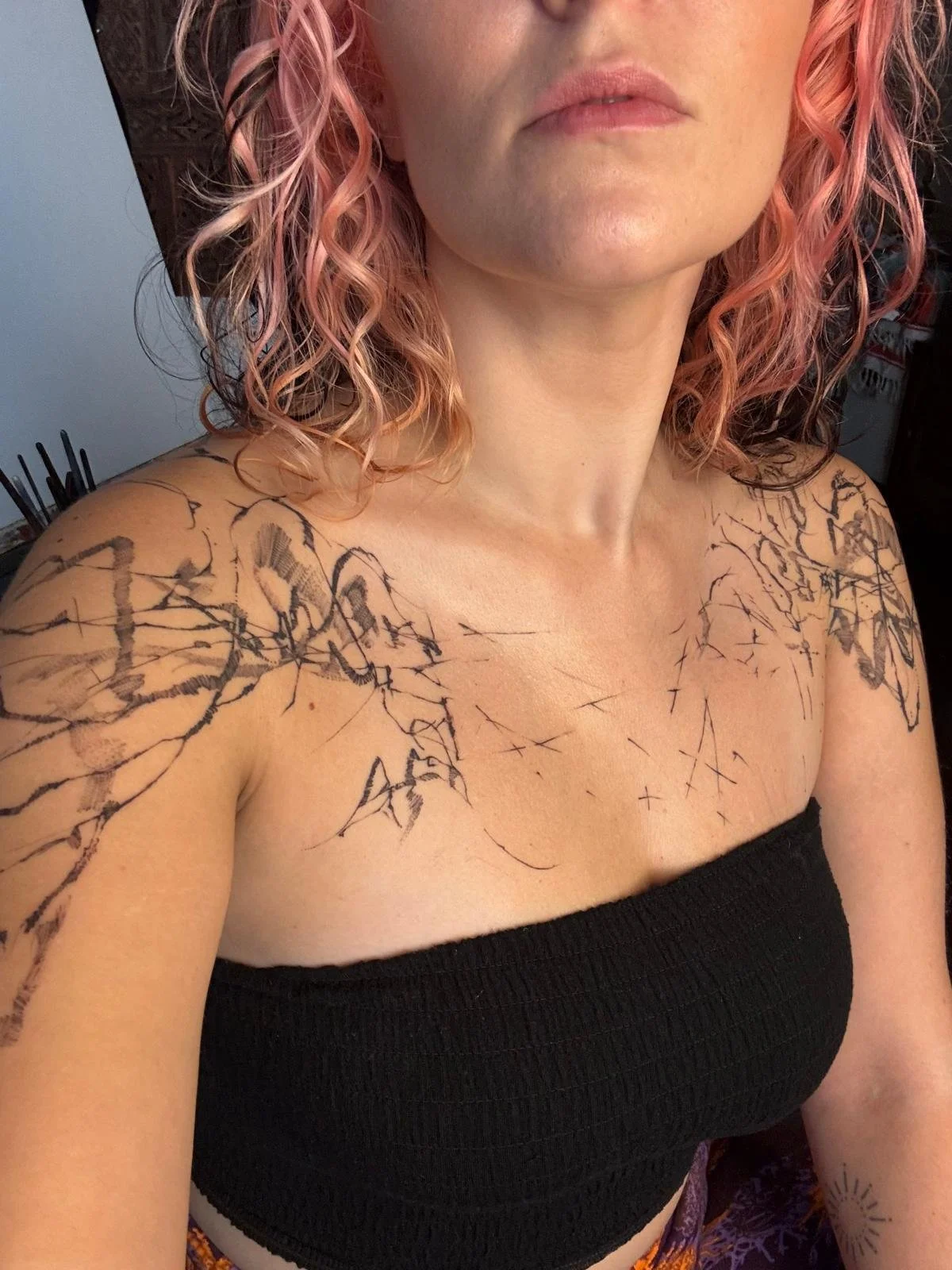 Close-up photo of a woman with curly pink hair, fair skin, and minimal makeup, wearing a black strapless top and displaying a large, intricate black tattoo design across her shoulders and upper chest.