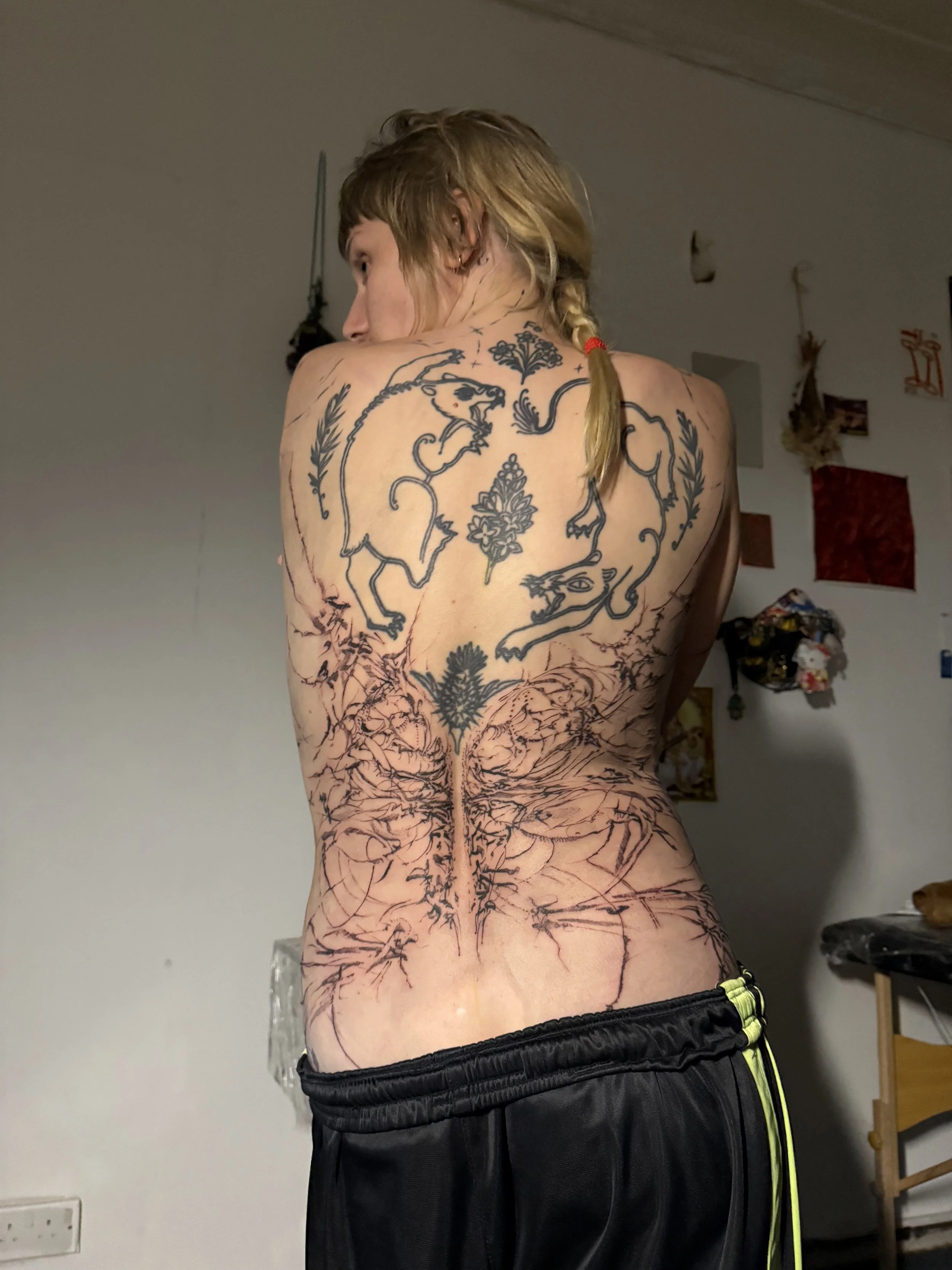 A person with long blonde hair in a braid, seen from the back, displaying large tattoos on their back and lower body. The tattoos feature a design with mythical creatures, flowers, and tree roots.