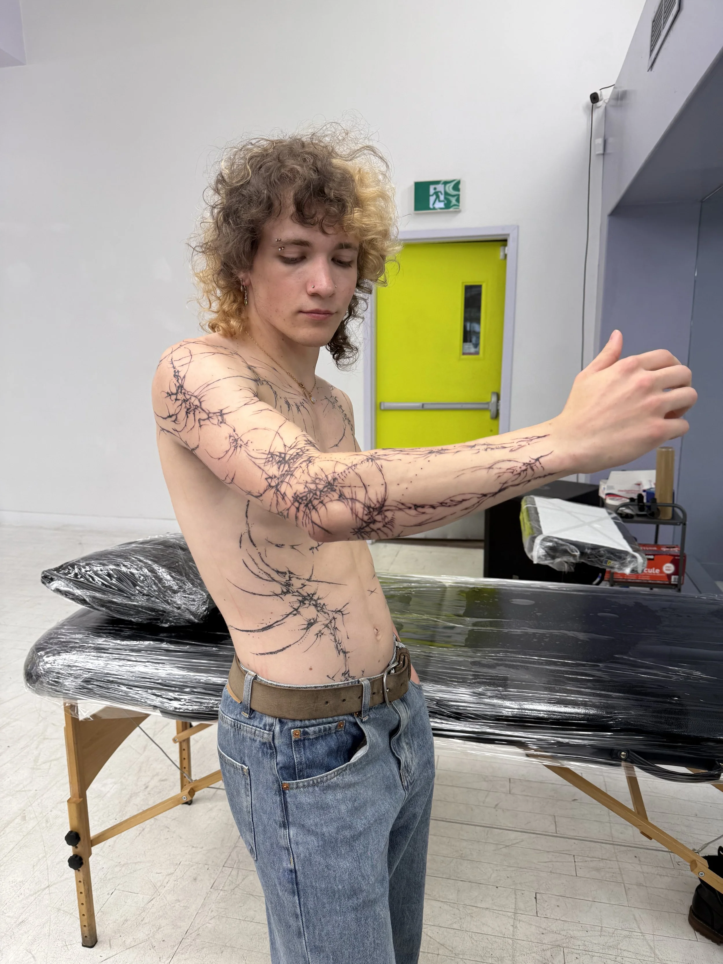 A shirtless young person with curly hair and tattoos on their arm and torso standing in a room with white walls, a yellow door, and a table covered with plastic wrap.