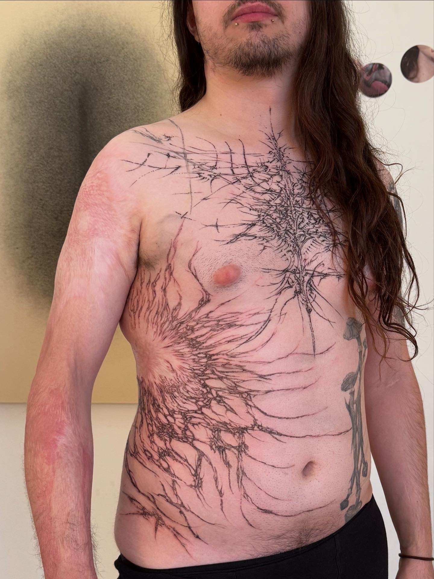 A person with long hair, facial piercings, and tattoos on their chest, arm, and torso. The tattoos are detailed black line art resembling a tree or branch pattern.