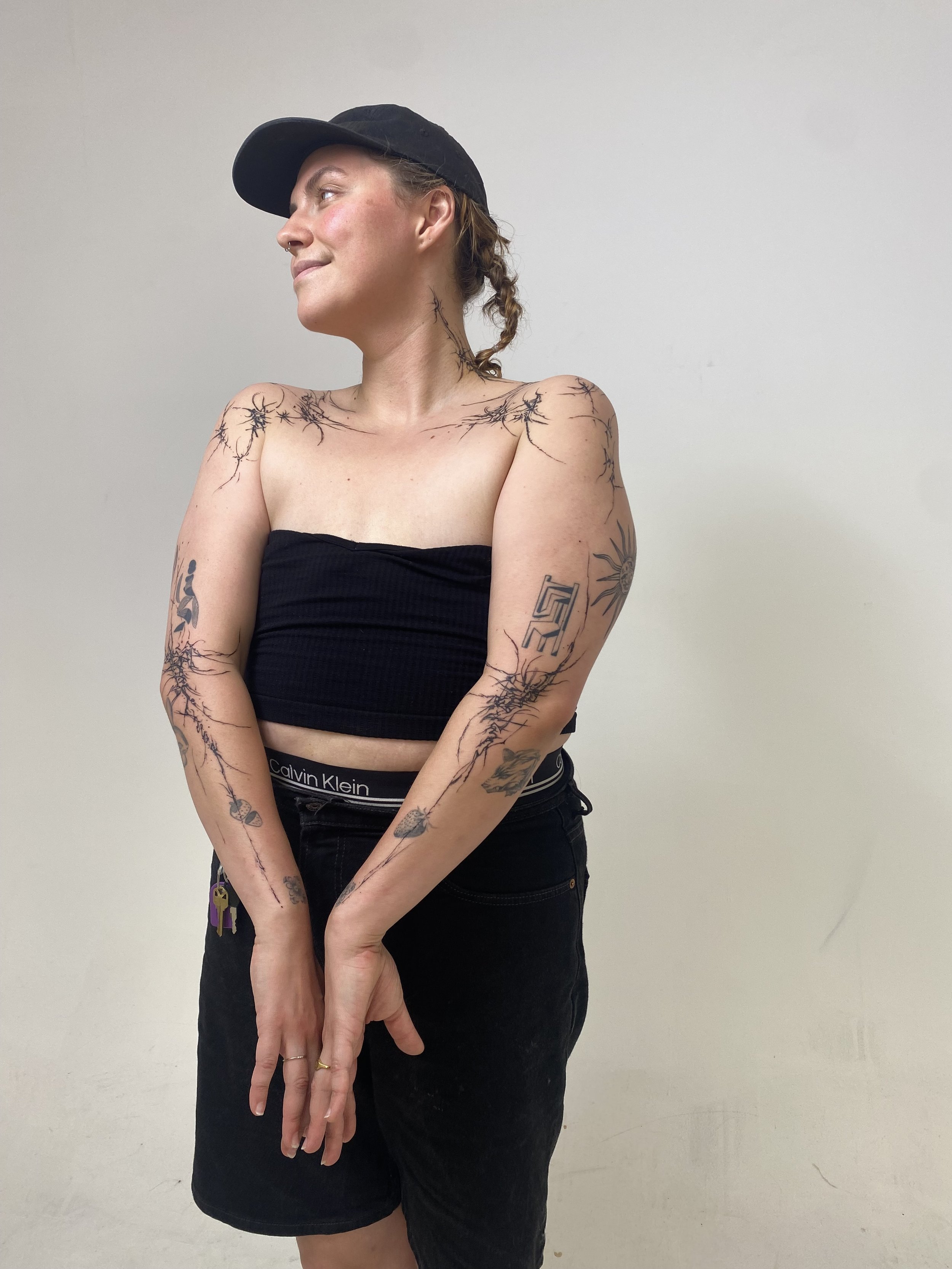 Tattooed woman with a black cap, black tube top, and black pants standing against a plain white wall, smiling and looking to her left.