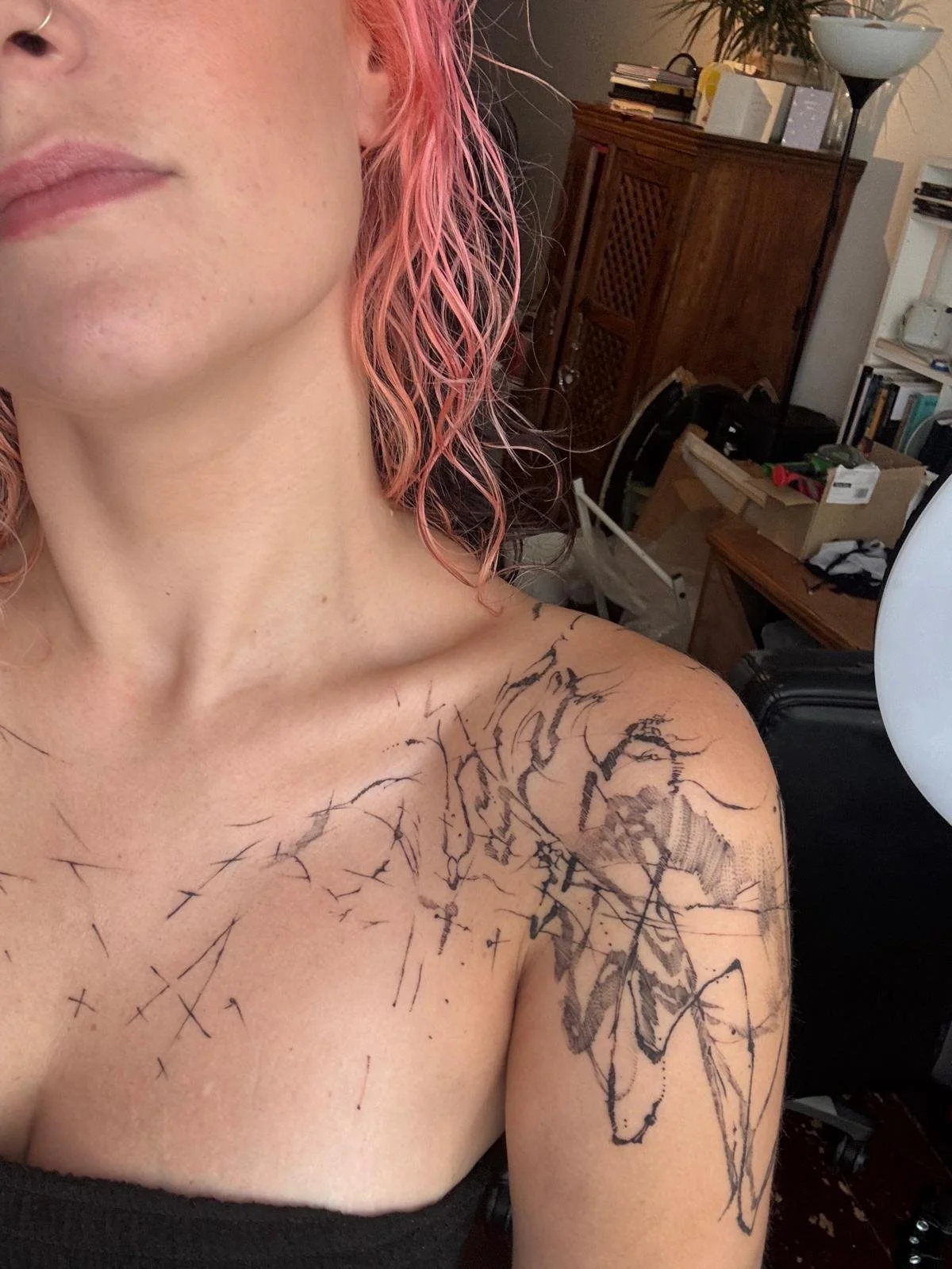A woman with pink hair shows a new abstract black ink tattoo on her shoulder and upper arm, in a room with shelves, boxes, and a lamp.