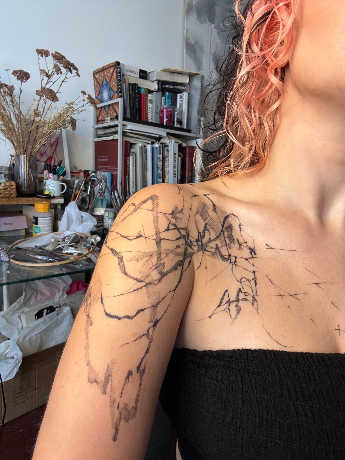 A woman with pink and black hair showing her shoulder and upper arm. She has a large abstract black tattoo on her shoulder and chest. The background shows a cluttered room with shelves, books, and art supplies.