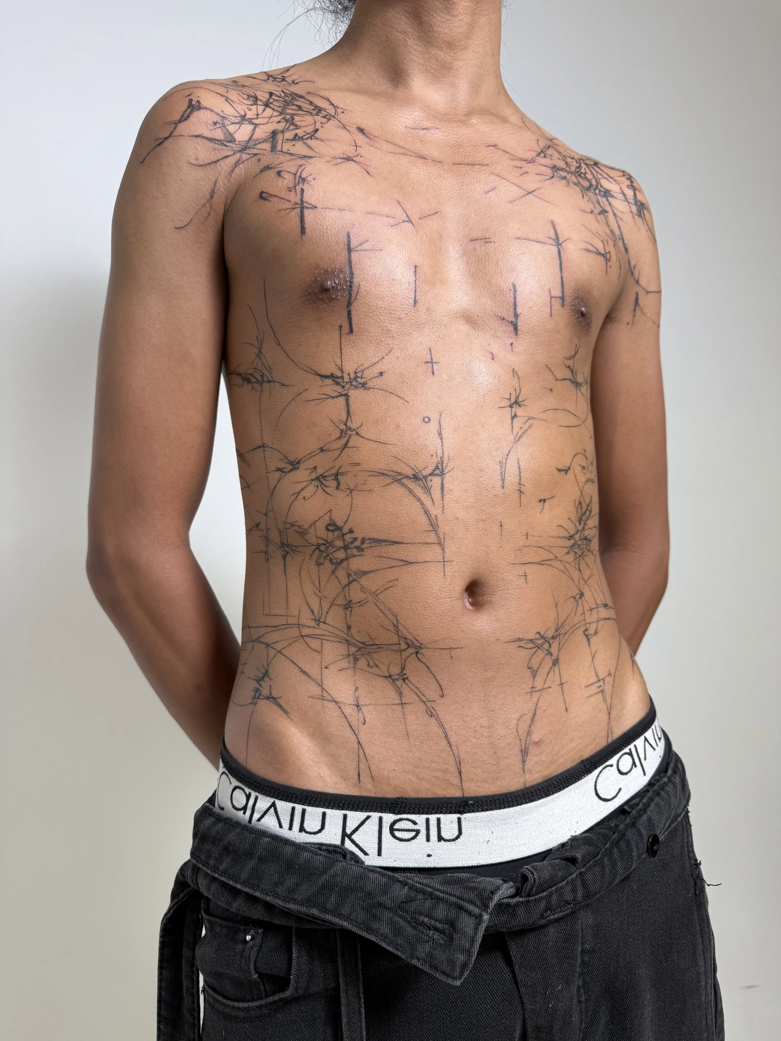 A person with a slender build, standing shirtless against a plain background, with numerous black ink tattoos of abstract, branching lines covering their shoulders, chest, and abdomen, and wearing black pants with the waistband of Calvin Klein underw