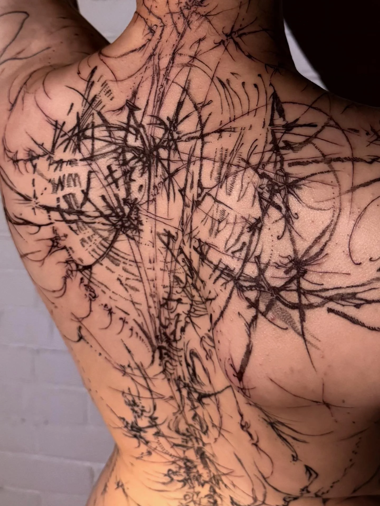 A close-up of a person's chest and shoulder covered in black ink tattoo lines that are abstract and chaotic in design.