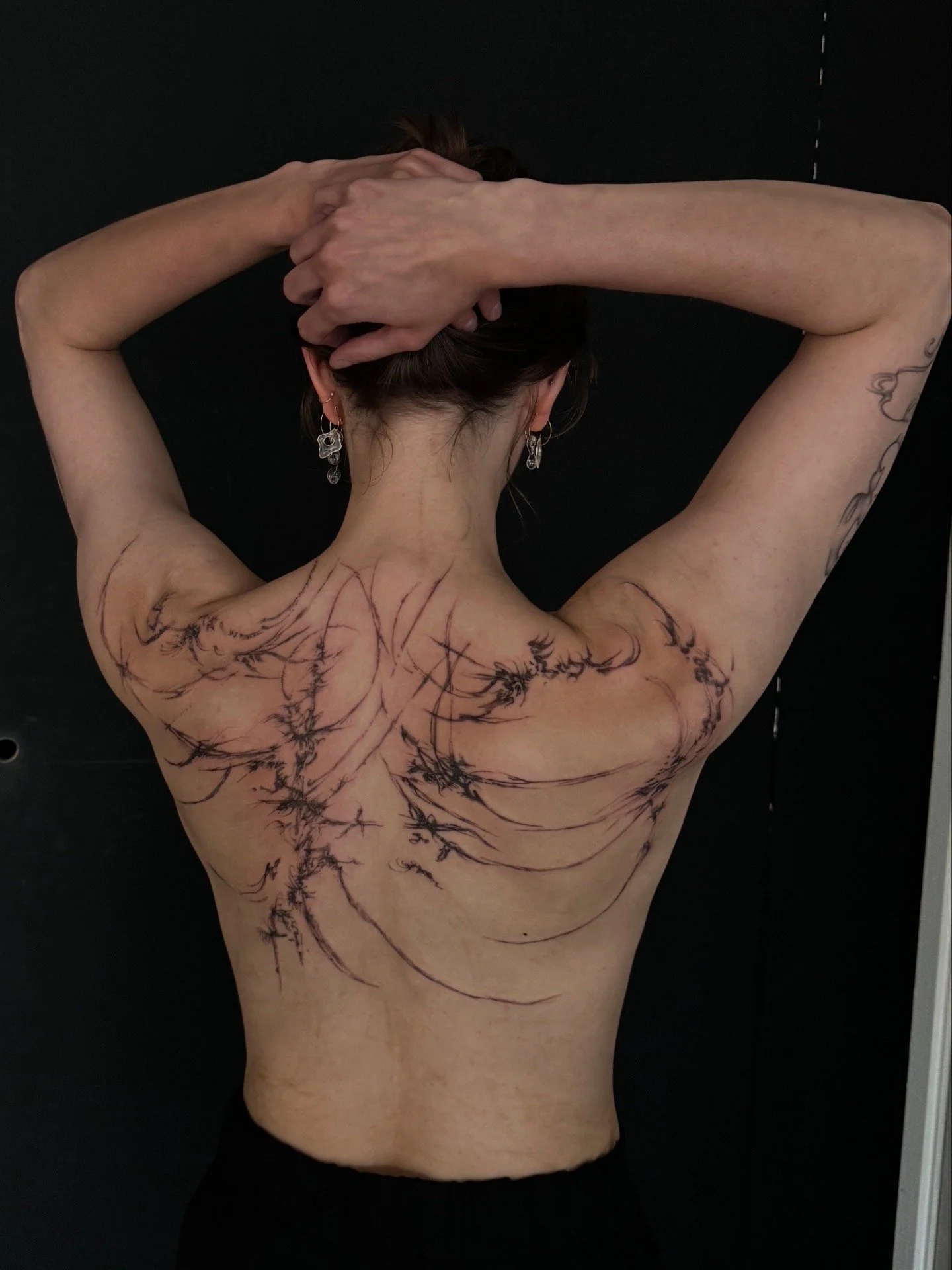 Back of a woman with multiple body scars and tattoos, arms raised with hands behind her head, earrings visible, dark background.
