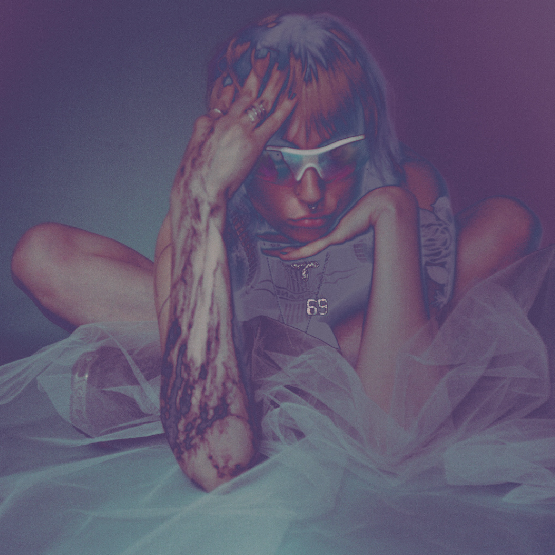A person with noise-esque abstract tattoos, wearing sunglasses, and jewelry, sitting on light-colored tulle fabric, with a thoughtful pose, looking downward.