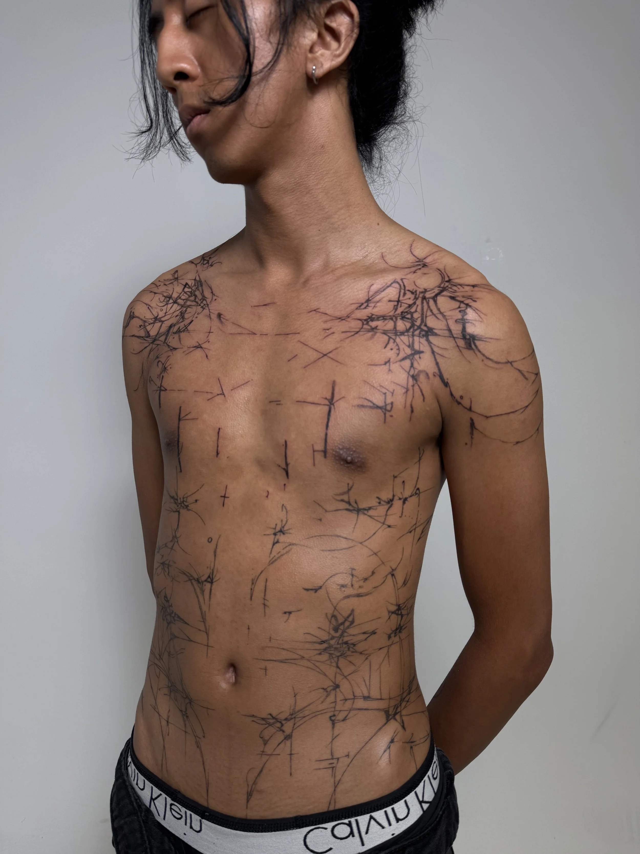 Person with black hair and earrings, shirtless, showing black tattoos that resemble barbed wire or thorny branches covering the shoulders, chest, and stomach, against a plain background.