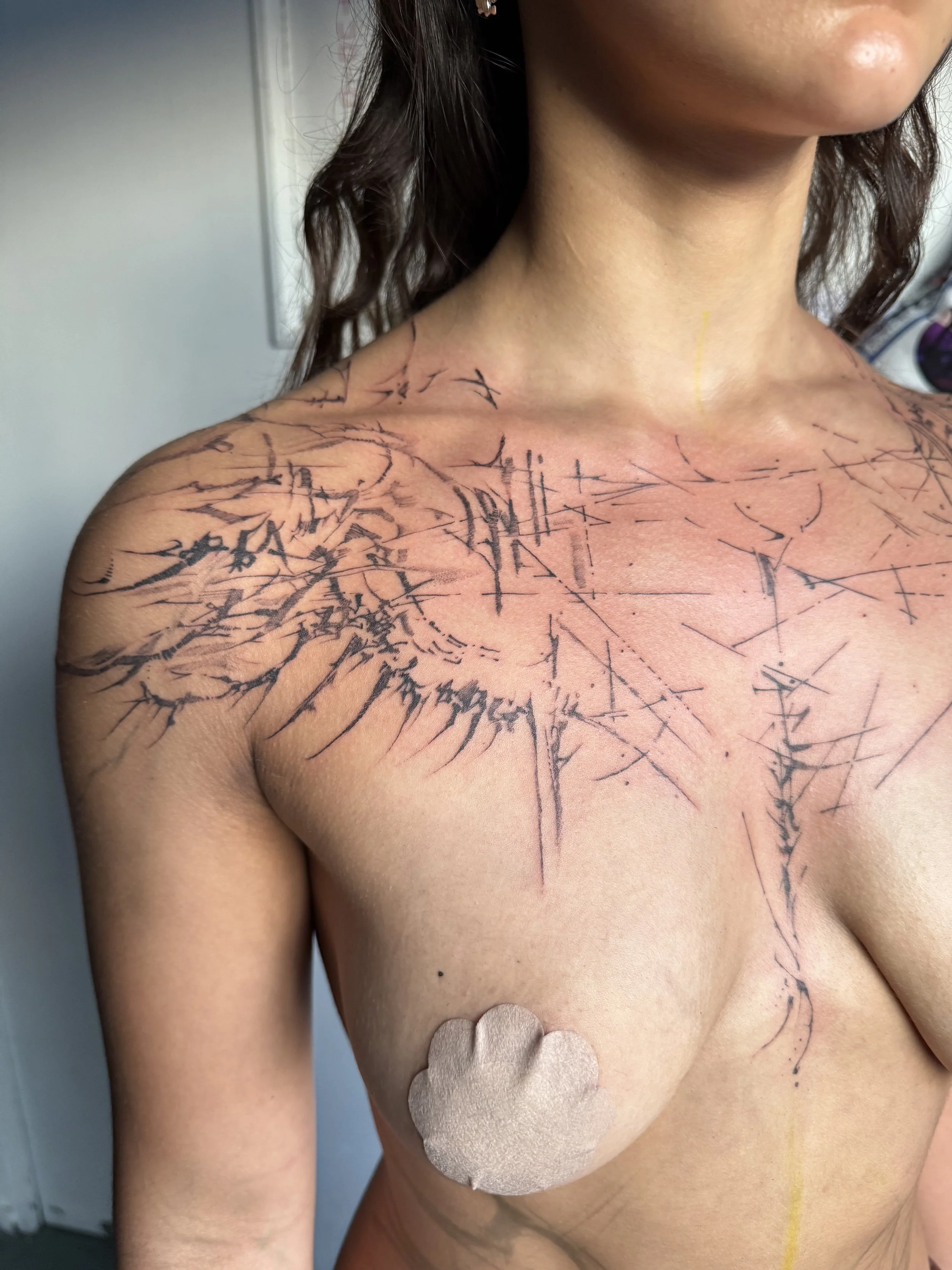 A woman with dark, curly hair and light skin has a recent tattoo on her shoulder and chest, consisting of black abstract lines and shapes that resemble a shattered glass or spider web pattern. She has a protective covering on her nipple.