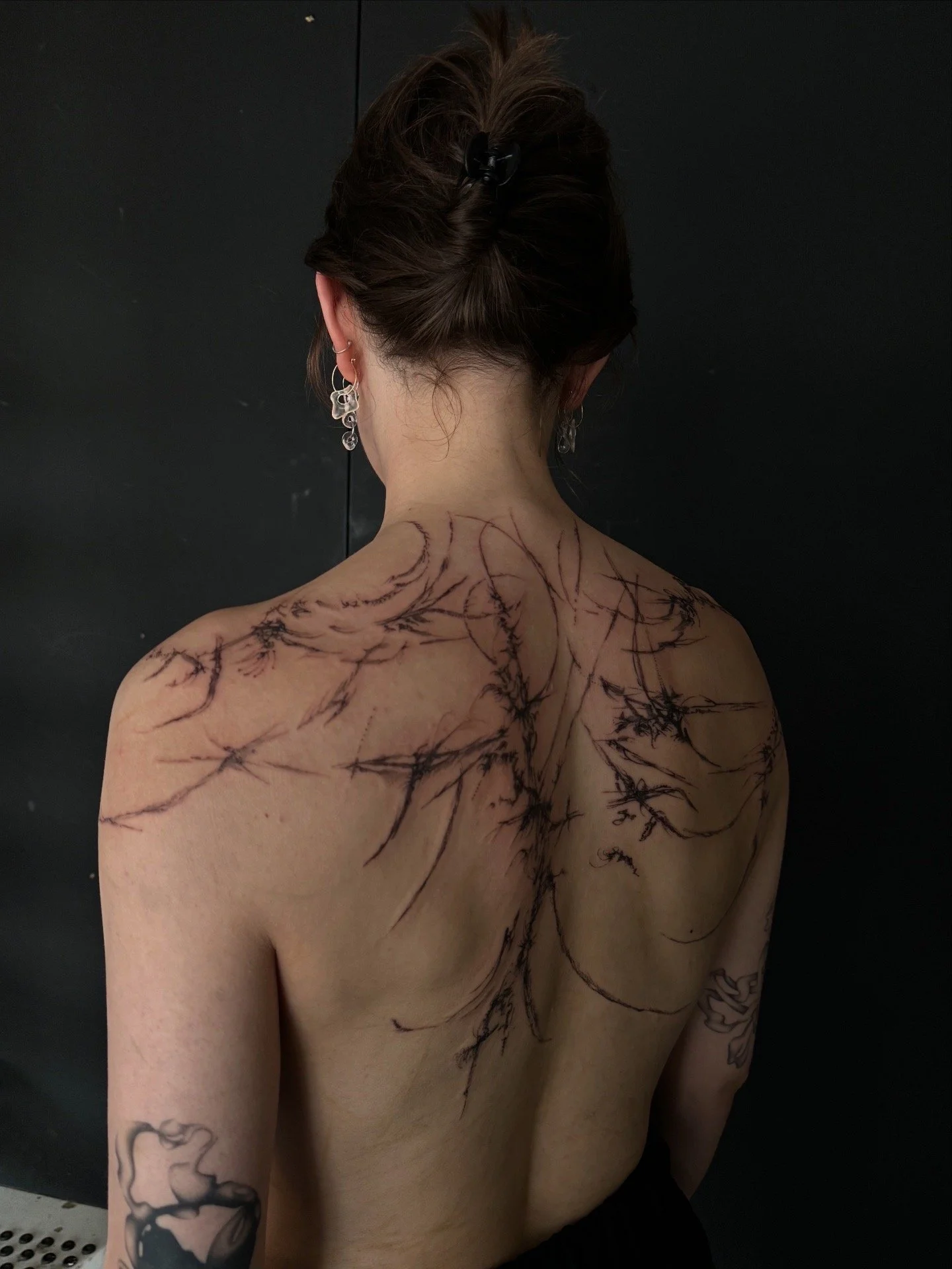 Back of a woman with dark hair in a bun, wearing earrings, with a large black ink tattoo of a tree or branch across her back and shoulders, against a black background.