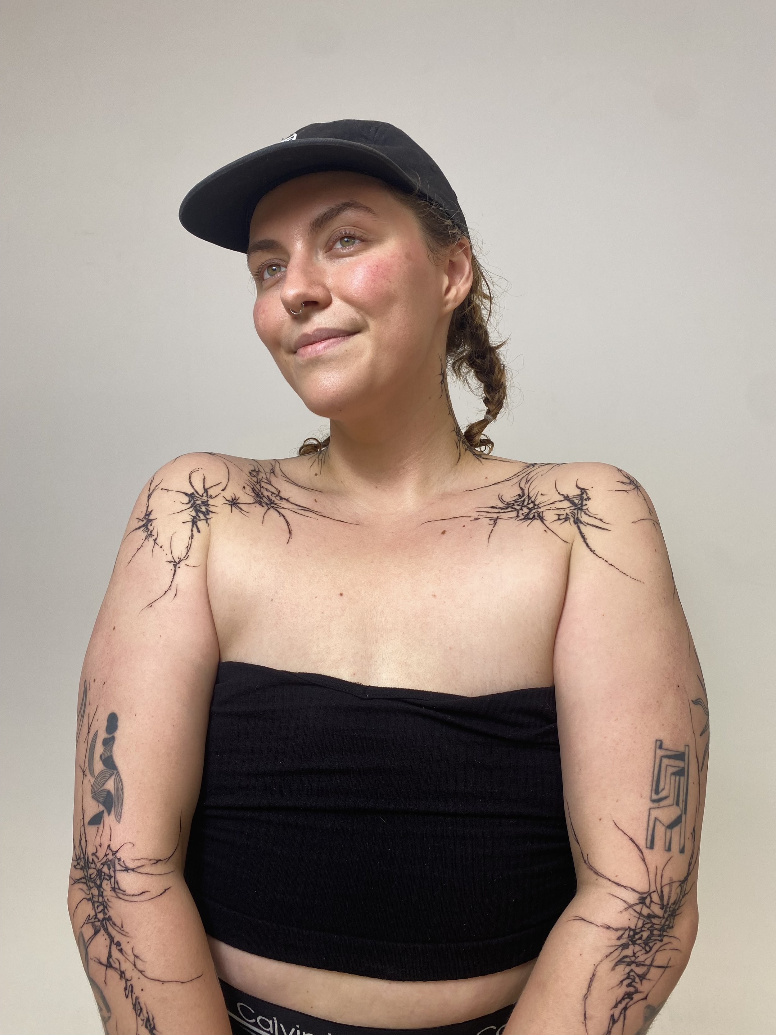 A young woman with tattoos on her shoulders and arms, wearing a black strapless top and a black baseball cap, smiling slightly and looking to her left against a plain light-colored wall.
