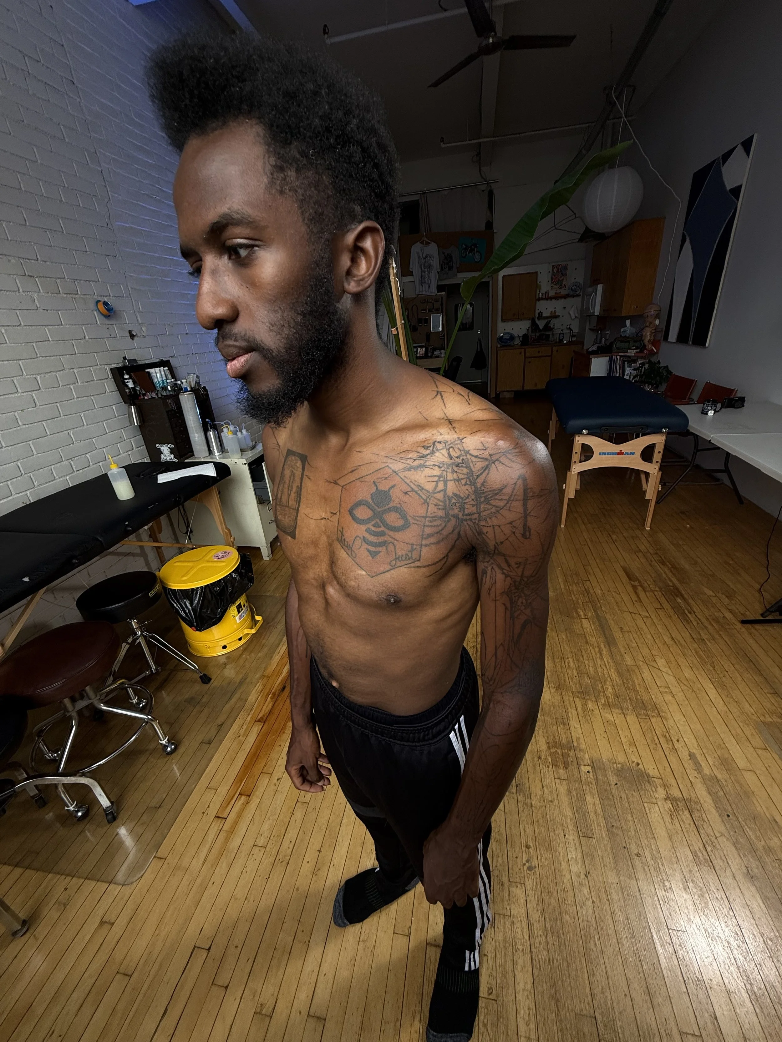 A shirtless man with tattoos on his chest and arms, standing in a room with wooden flooring and furniture, looking to his left.